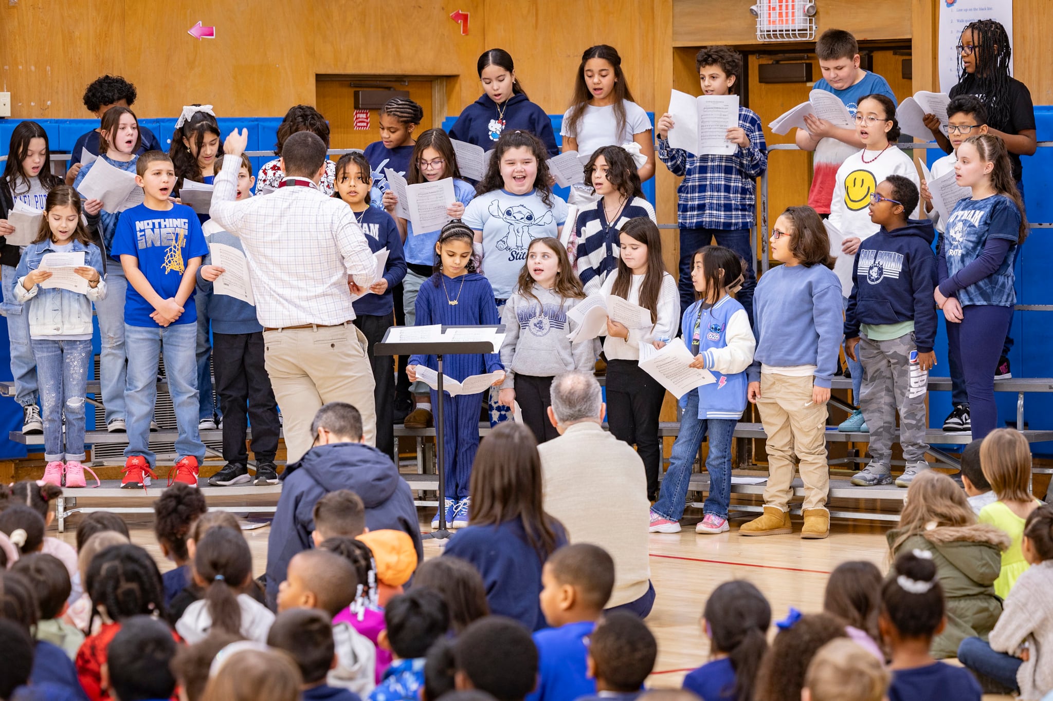 A photograph of a student choir on stage while a group of students sit on chairs in the foreground.