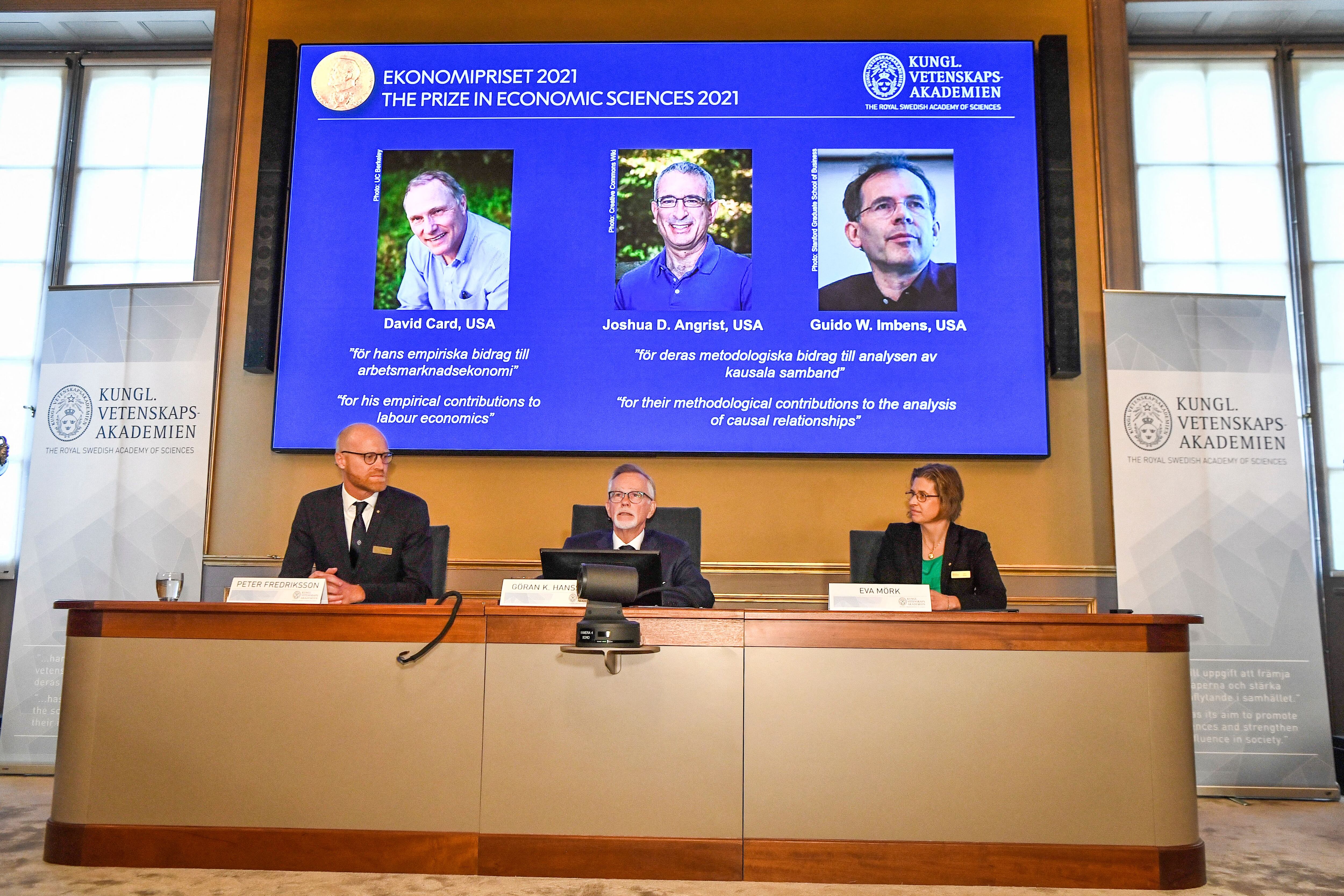 Three panelists of the Nobel Prize sit at a desk as a projector shows the winners of the Nobel Prize for Economics.
