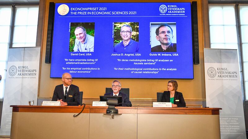 Three panelists of the Nobel Prize sit at a desk as a projector shows the winners of the Nobel Prize for Economics.