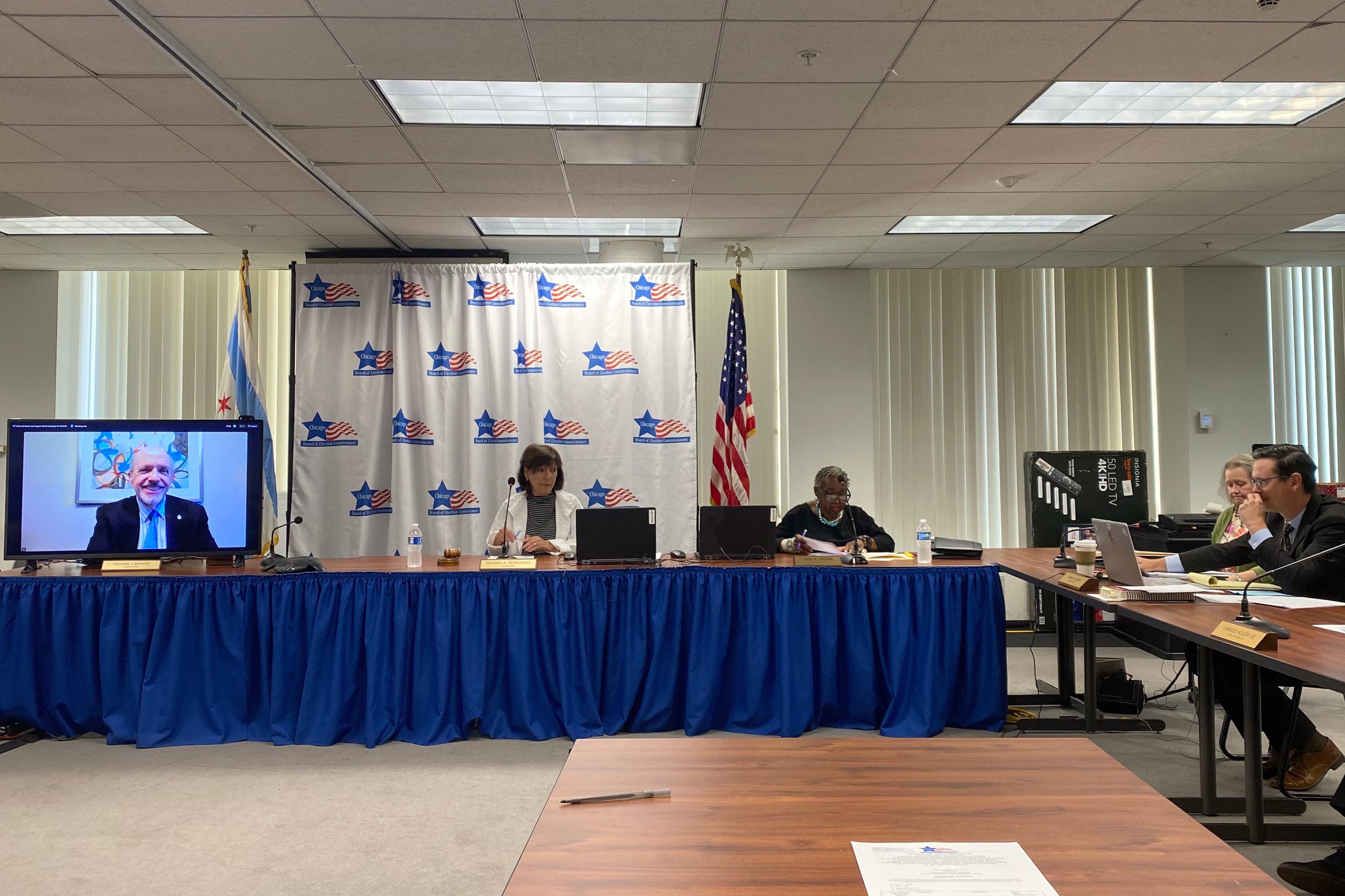 A small group of people sit around tables in a meeting room with a large video screen on the side and flags and curtains in the background.