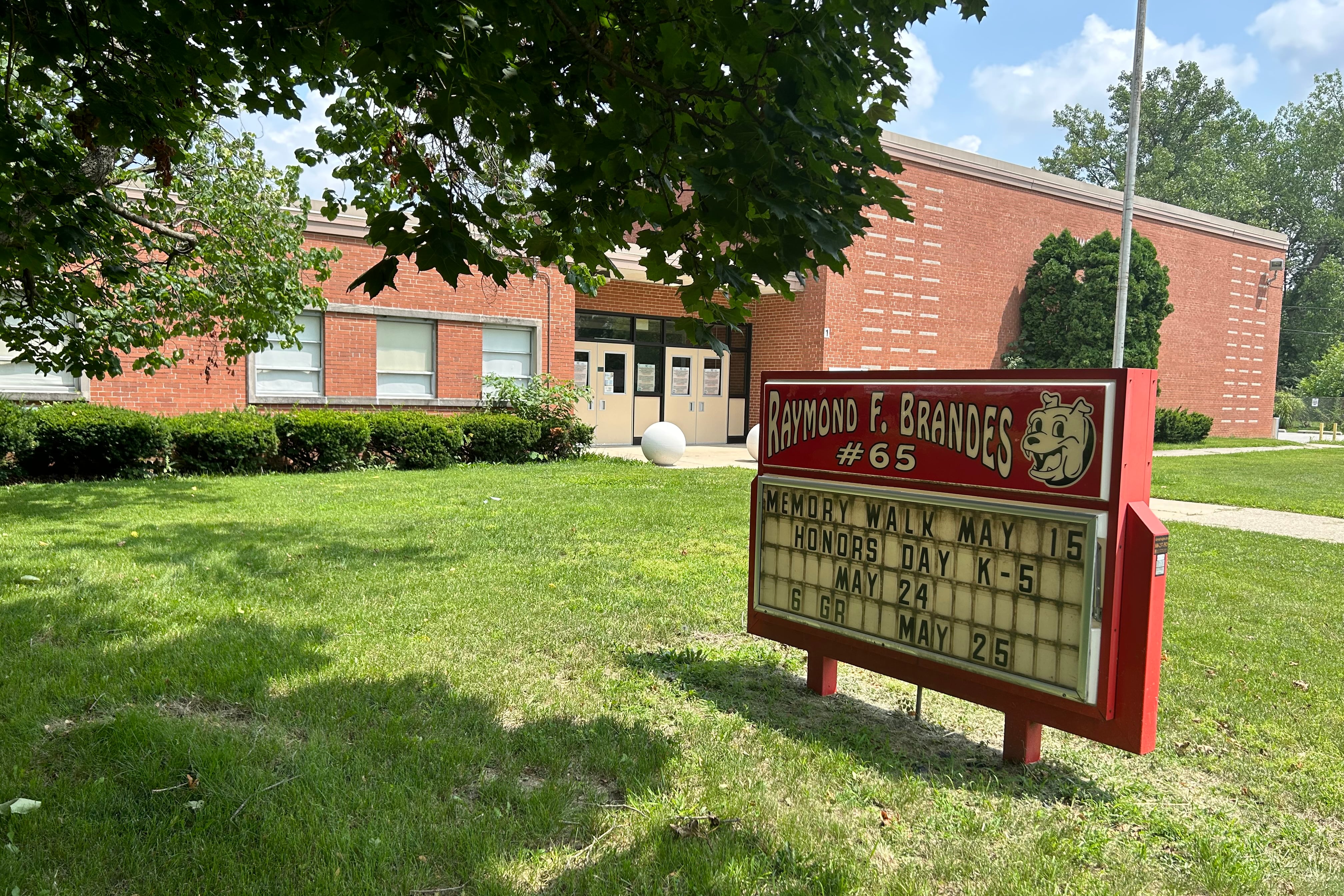 A sign reading “Raymond F. Brandes number 65” sits in a green lawn with a brick building in the background.