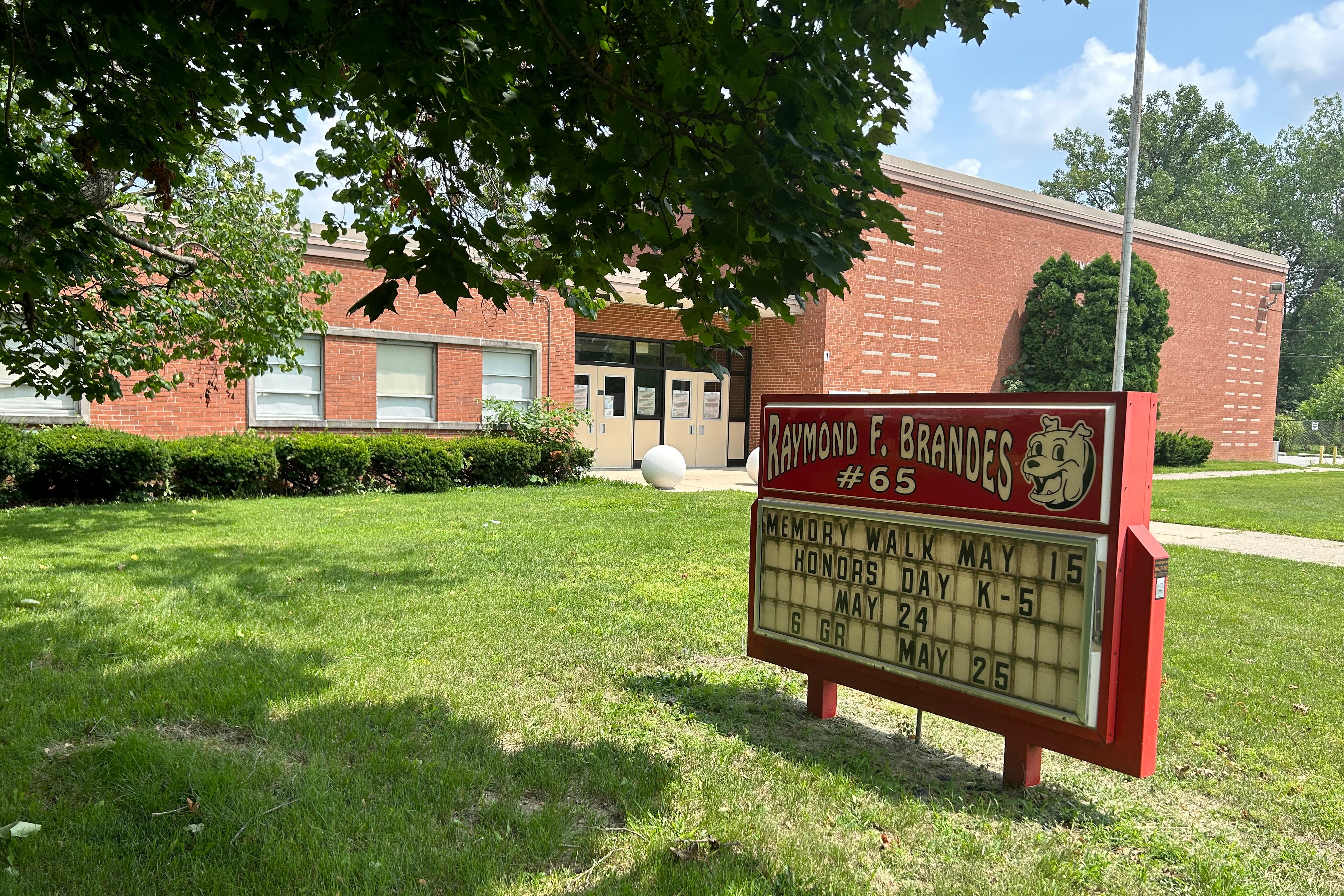 A sign reading “Raymond F. Brandes number 65” sits in a green lawn with a brick building in the background.