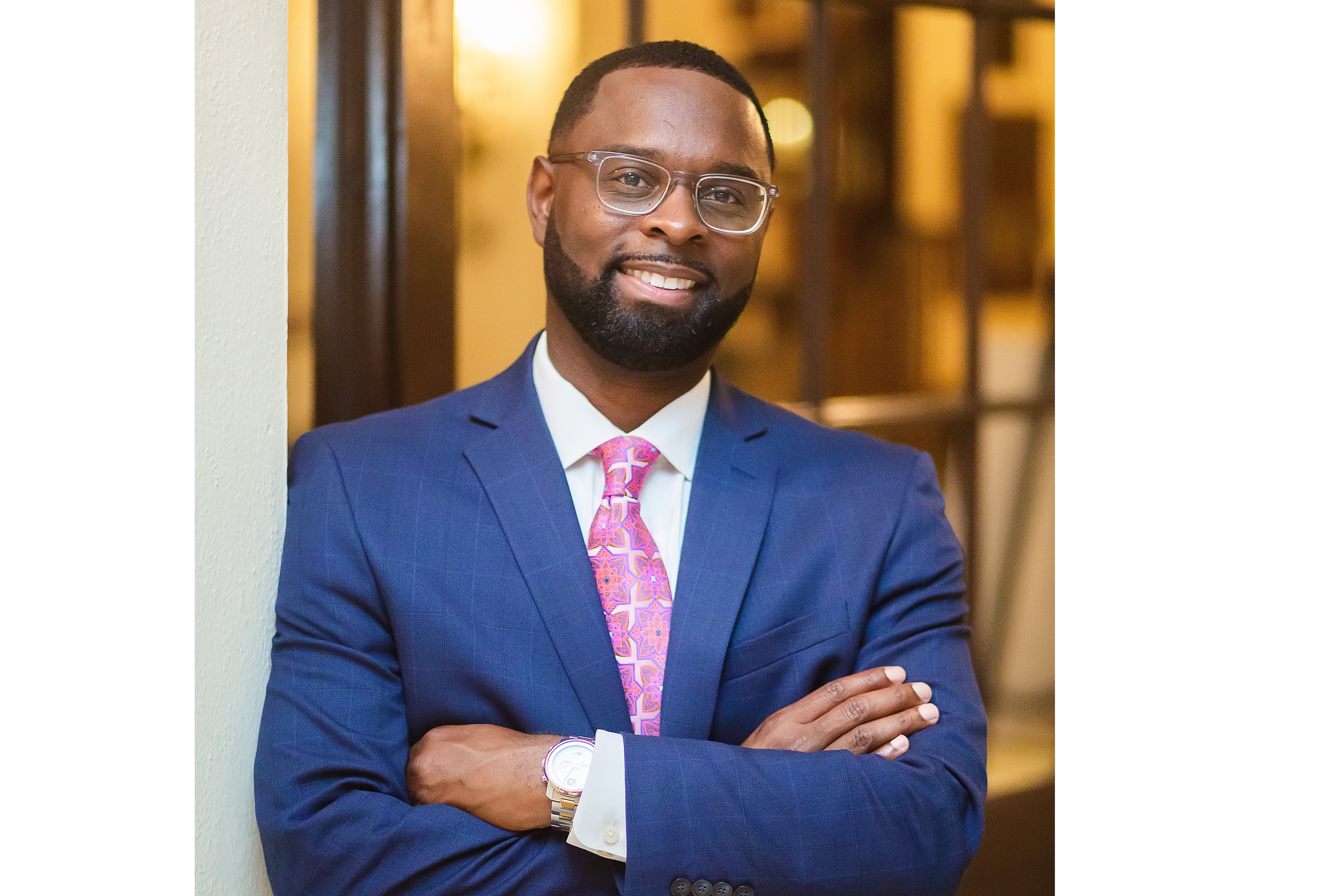 A man wearing glasses and a blue suit with a reddish-pink tie crosses his arms and smiles in a posed portrait.