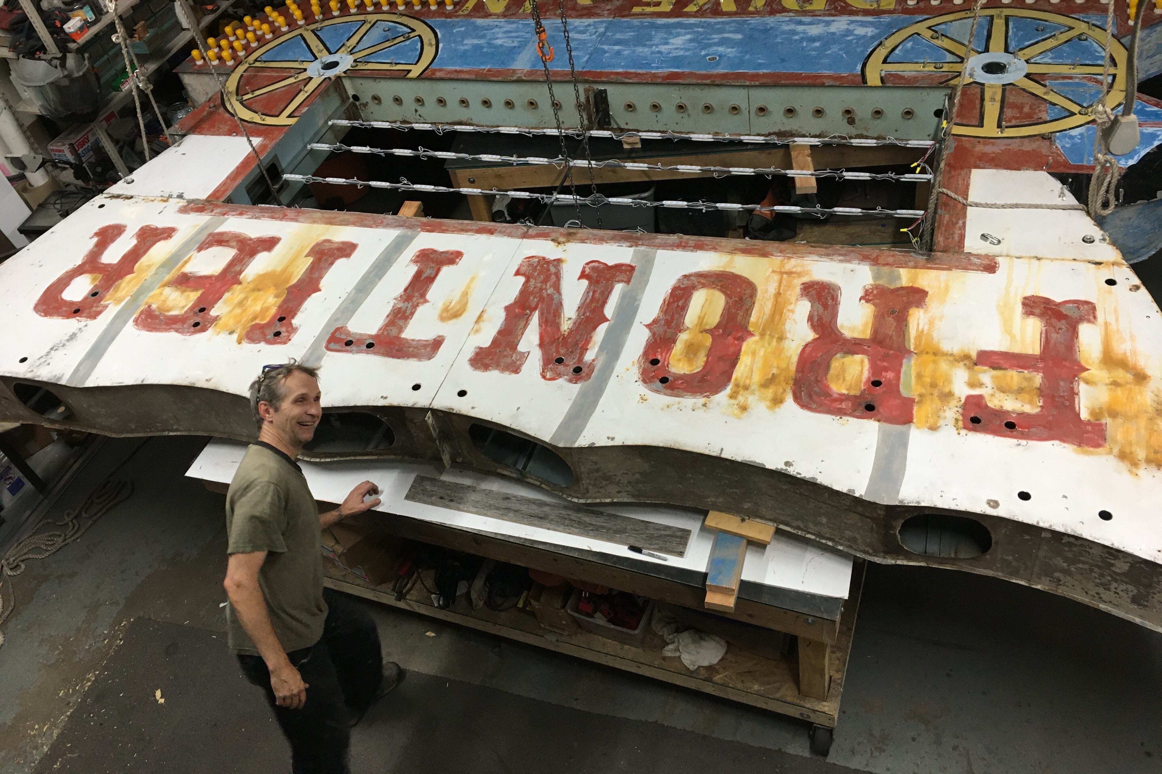 A man in a grey shirt stands in front of a giant sign that reads “frontier.”
