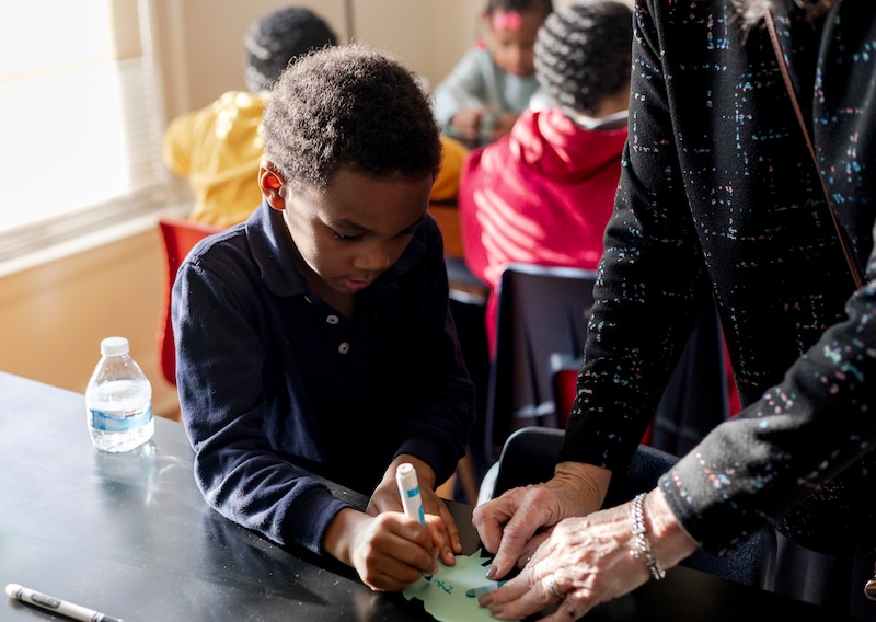 A young child draws at a table next to an adult in a house.