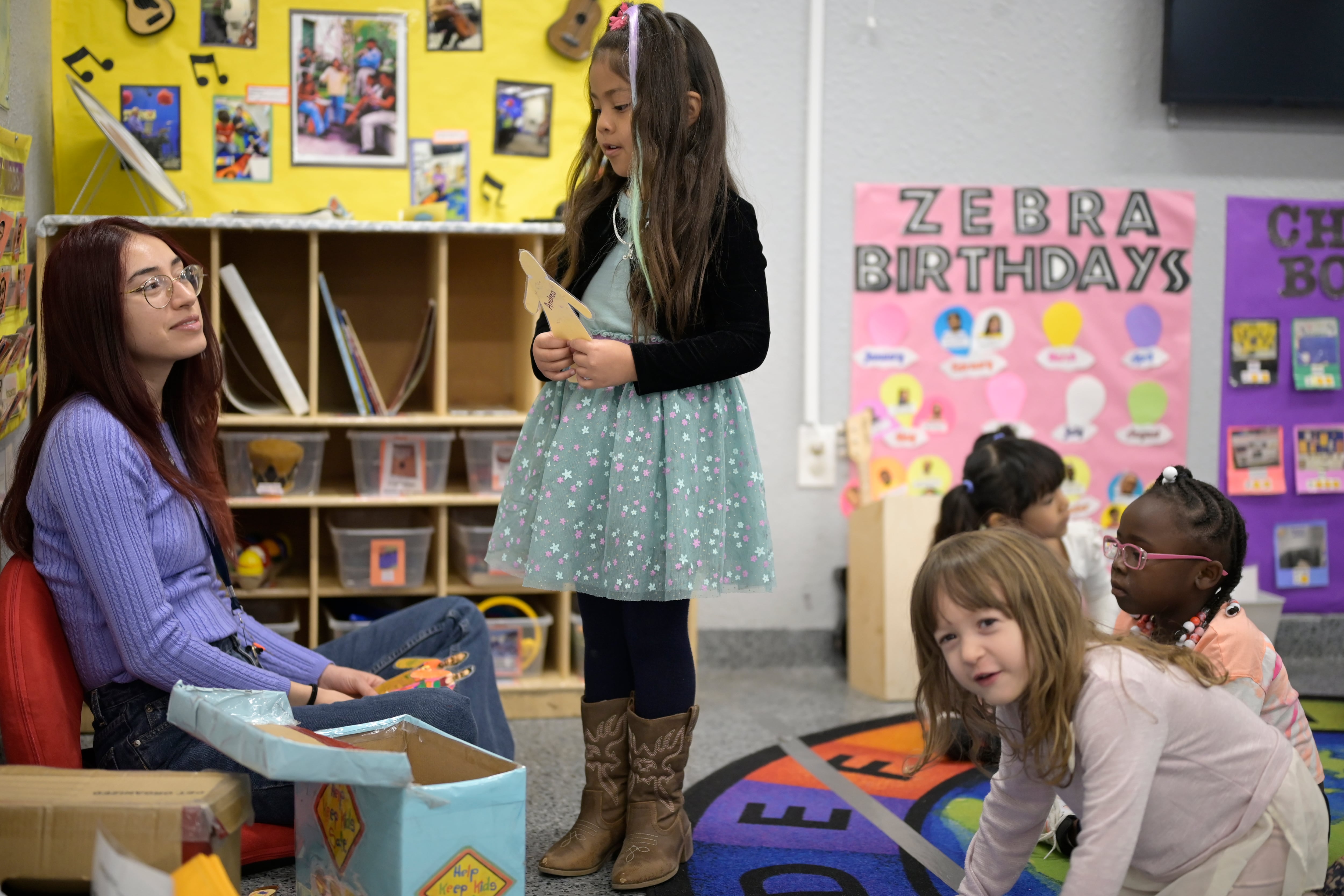 A young girl with long brown hair wearing a dress and cowboy boots is standing in front of a sitting teacher and there are other children on the classroom rug behind her.