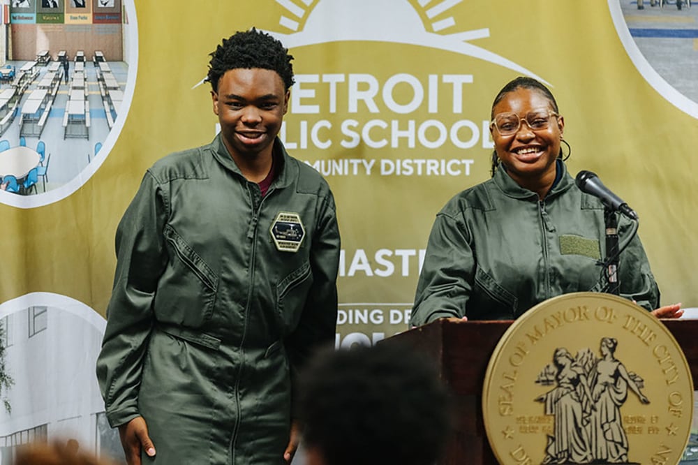 Two Black high school students smile as they speak from behind a wooden podium with a microphone and in front of a yellow banner. They are wearing green jumpsuits.