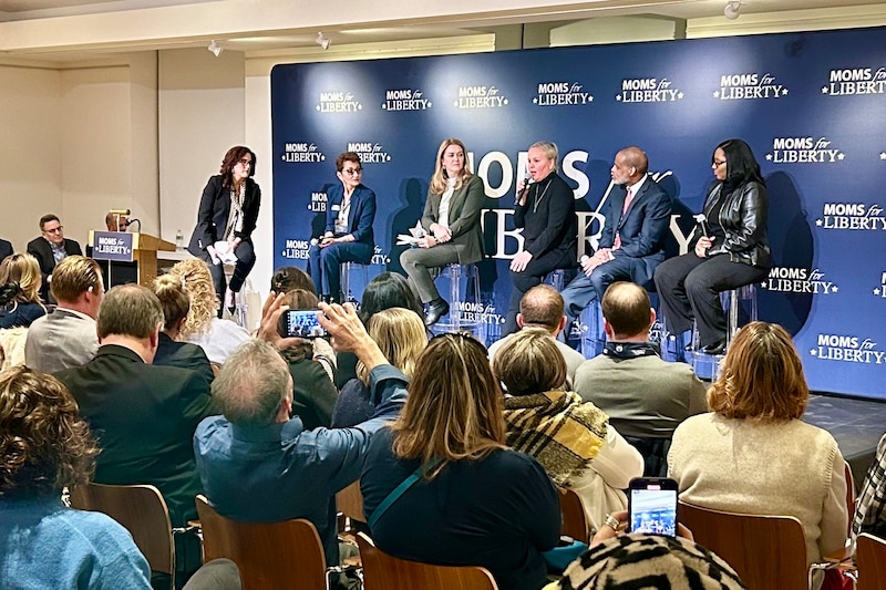 A group of men and women in suits sit on a stage in front of a small crowd.