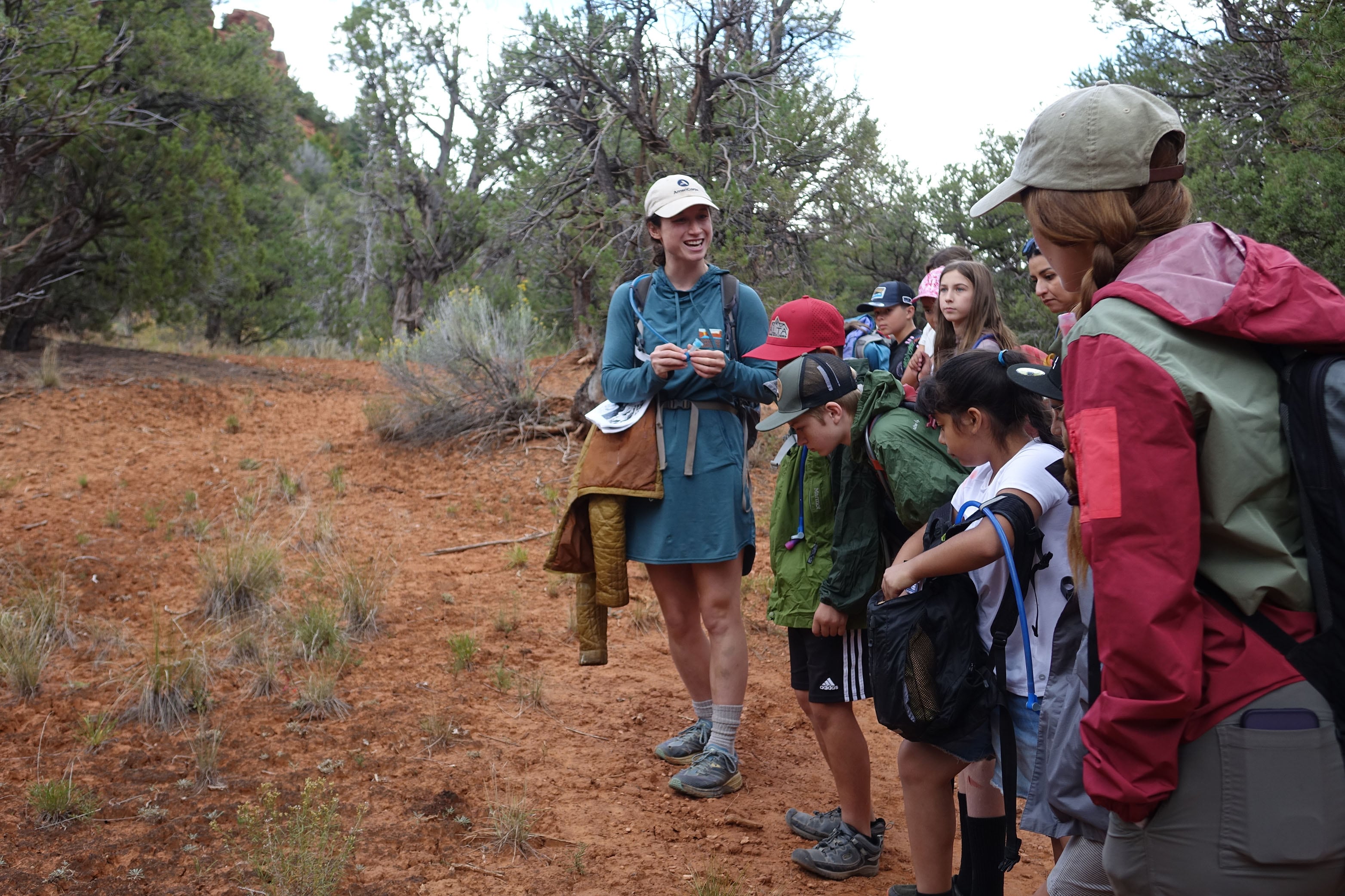 An adult woman stands next to a group of young students outside along a trail.