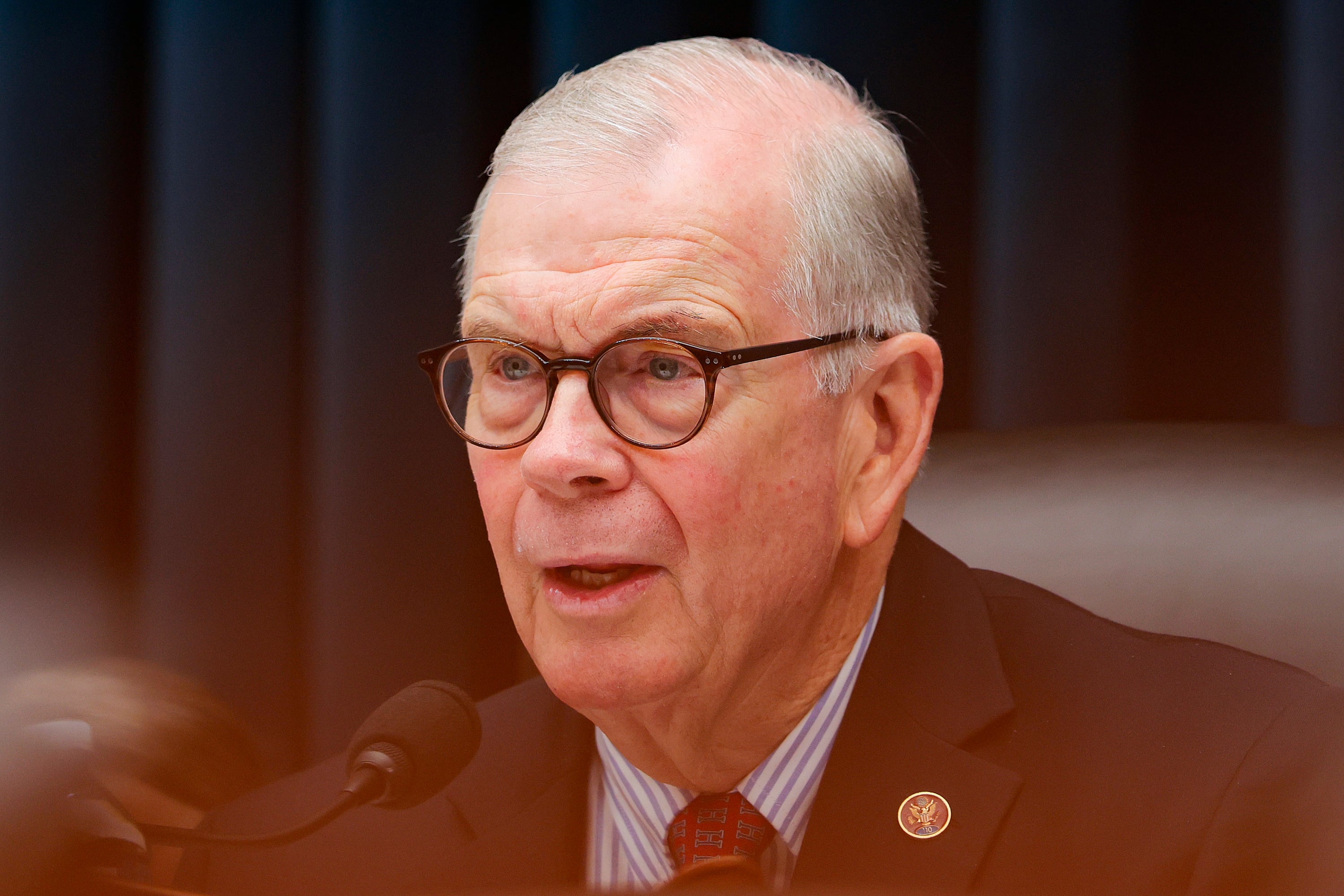 A photograph of a white male with glasses speaking into a microphone.
