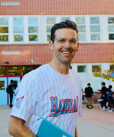 Headshot of man with brown hair standing in front of a school building. His shirt says, "Manual" on it.