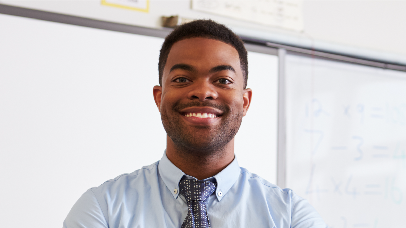 A teacher smiles and stands in front of a whiteboard in his classroom.