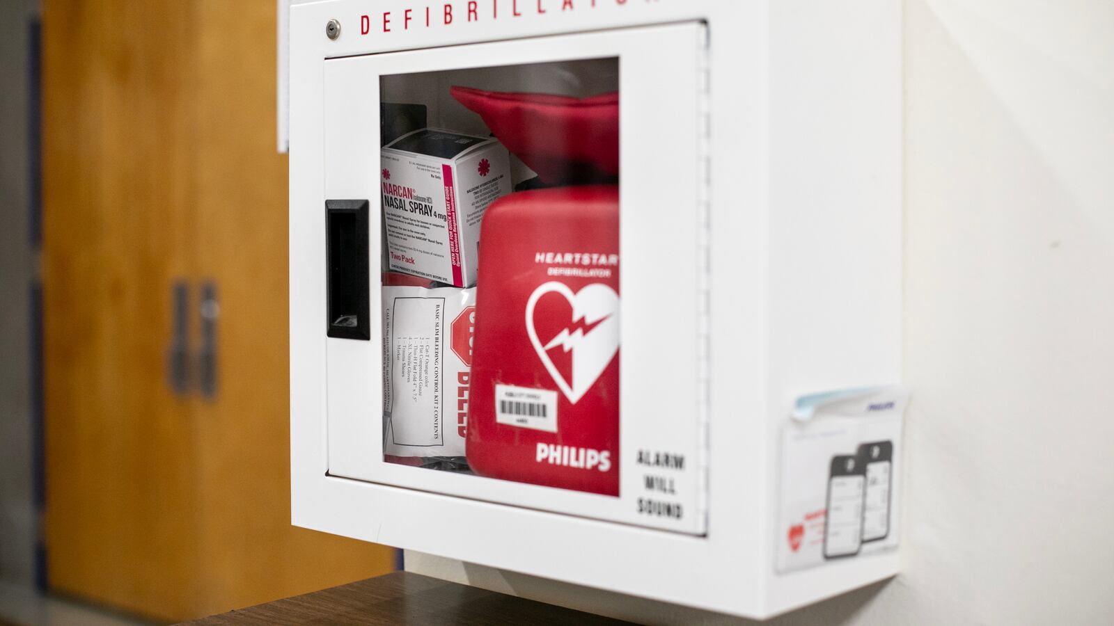 An emergency box mounted in the hallway of a building, containing a defibrillator and medications.