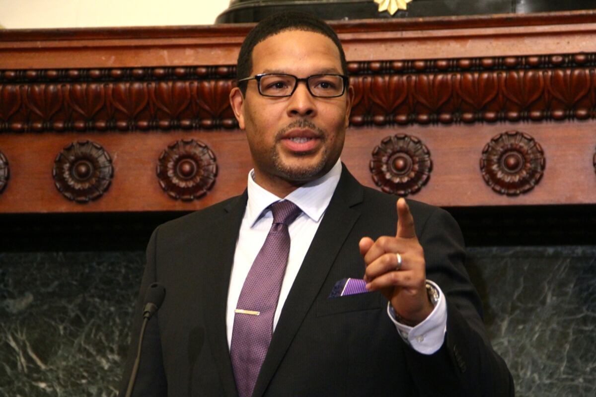 A man in a light shirt and dark suit with a purple pocket square and wearing glasses points his finger while giving a speech at a press conference.