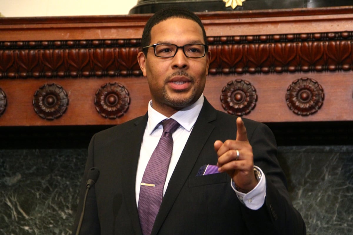 A man in a light shirt and dark suit with a purple pocket square and wearing glasses points his finger while giving a speech at a press conference.
