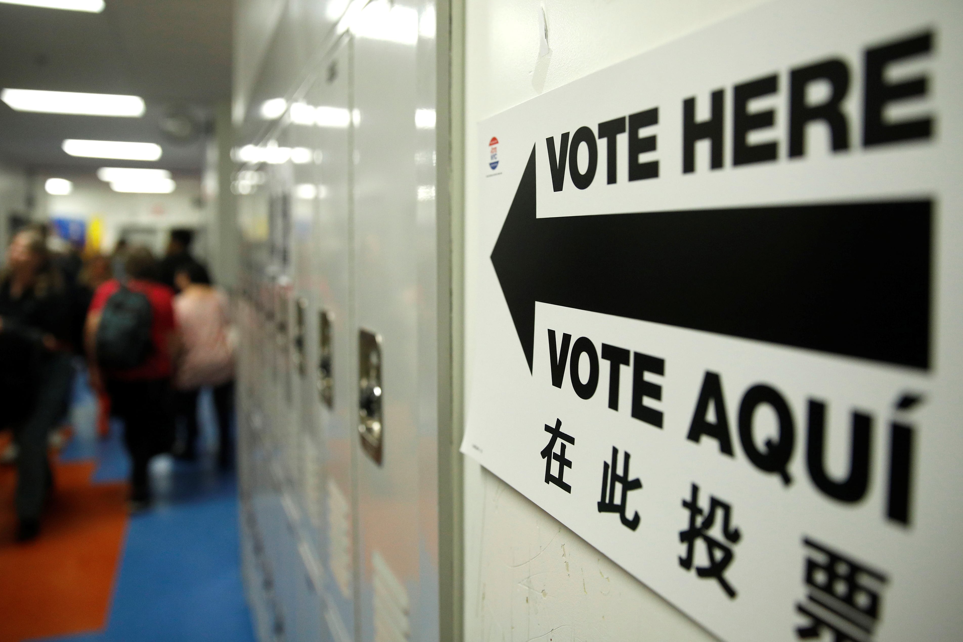 A sign that reads "VOTE HERE" in three different languages is posted on a wall near lockers and a group of people in the background in a school.