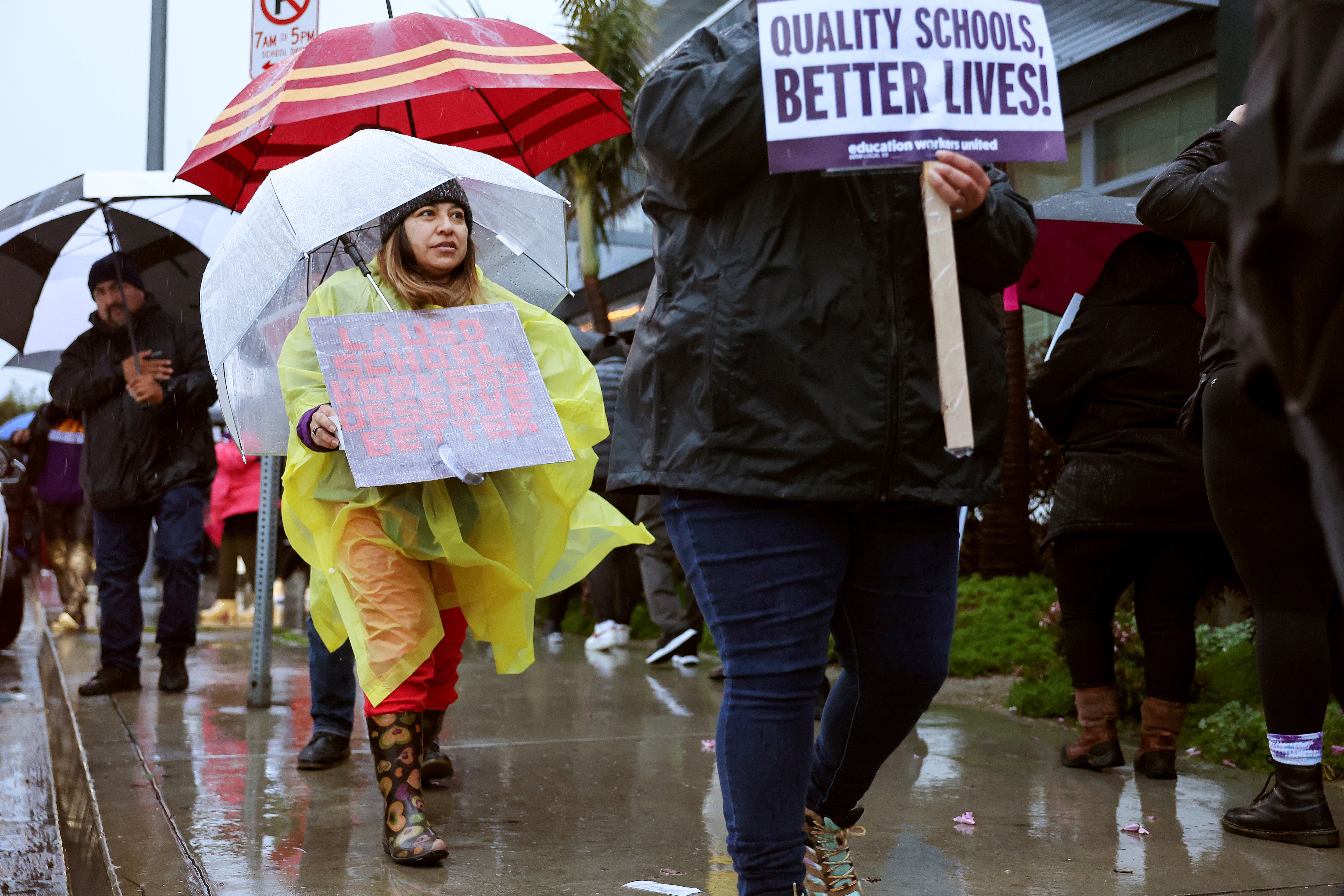 People in rain coats hold signs and umbrellas while walking outside during a strike.
