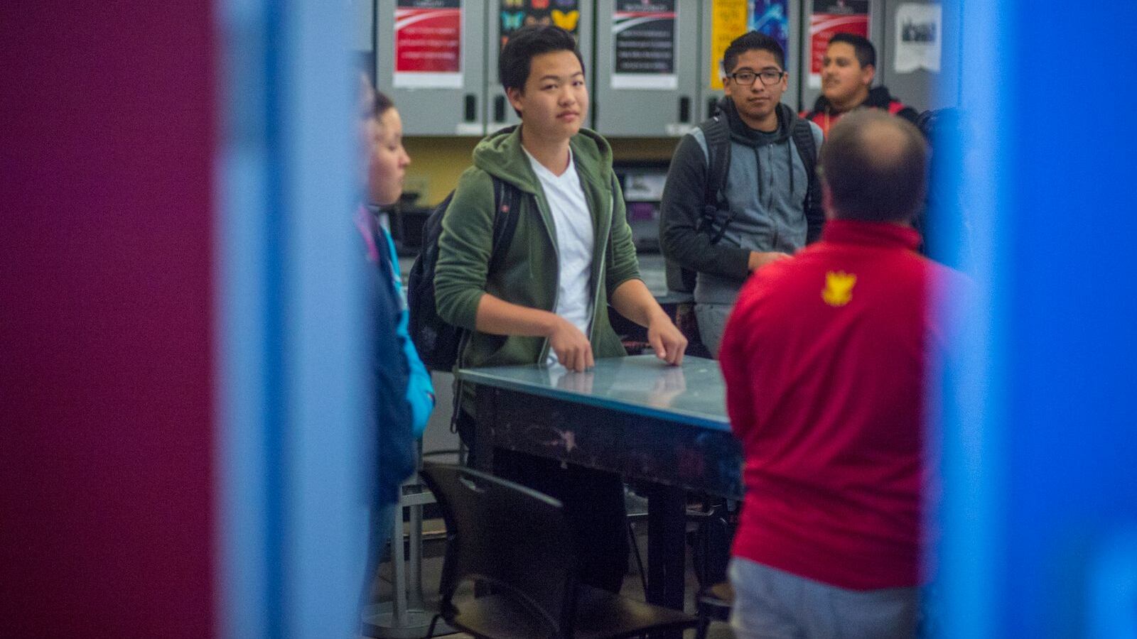 Four high school students stand around a table speaking with an older adult wearing a red jacket. They are seen through a blue doorframe.