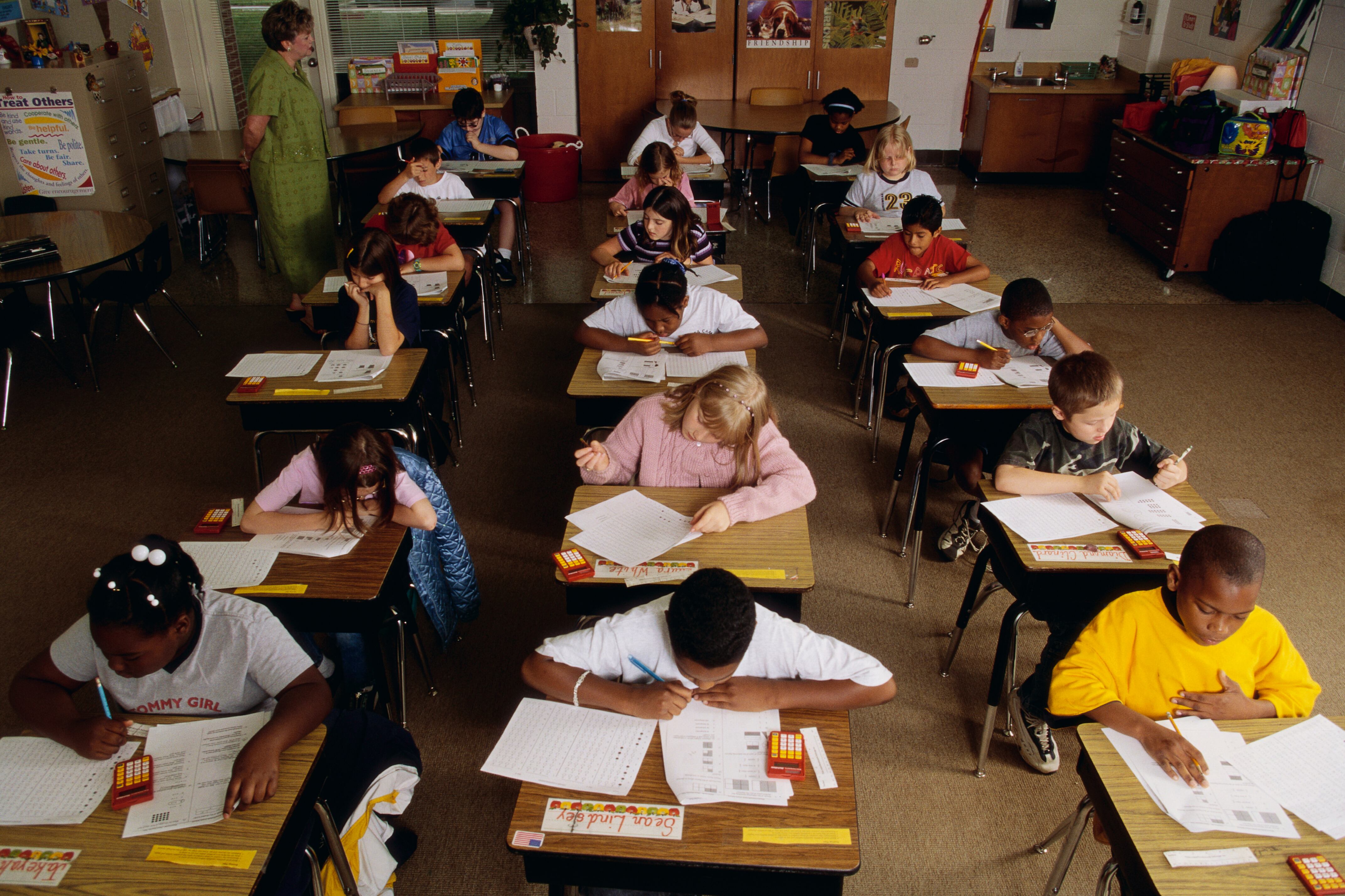 Eighteen elementary students take a standardized test at their desks while a teacher watches.