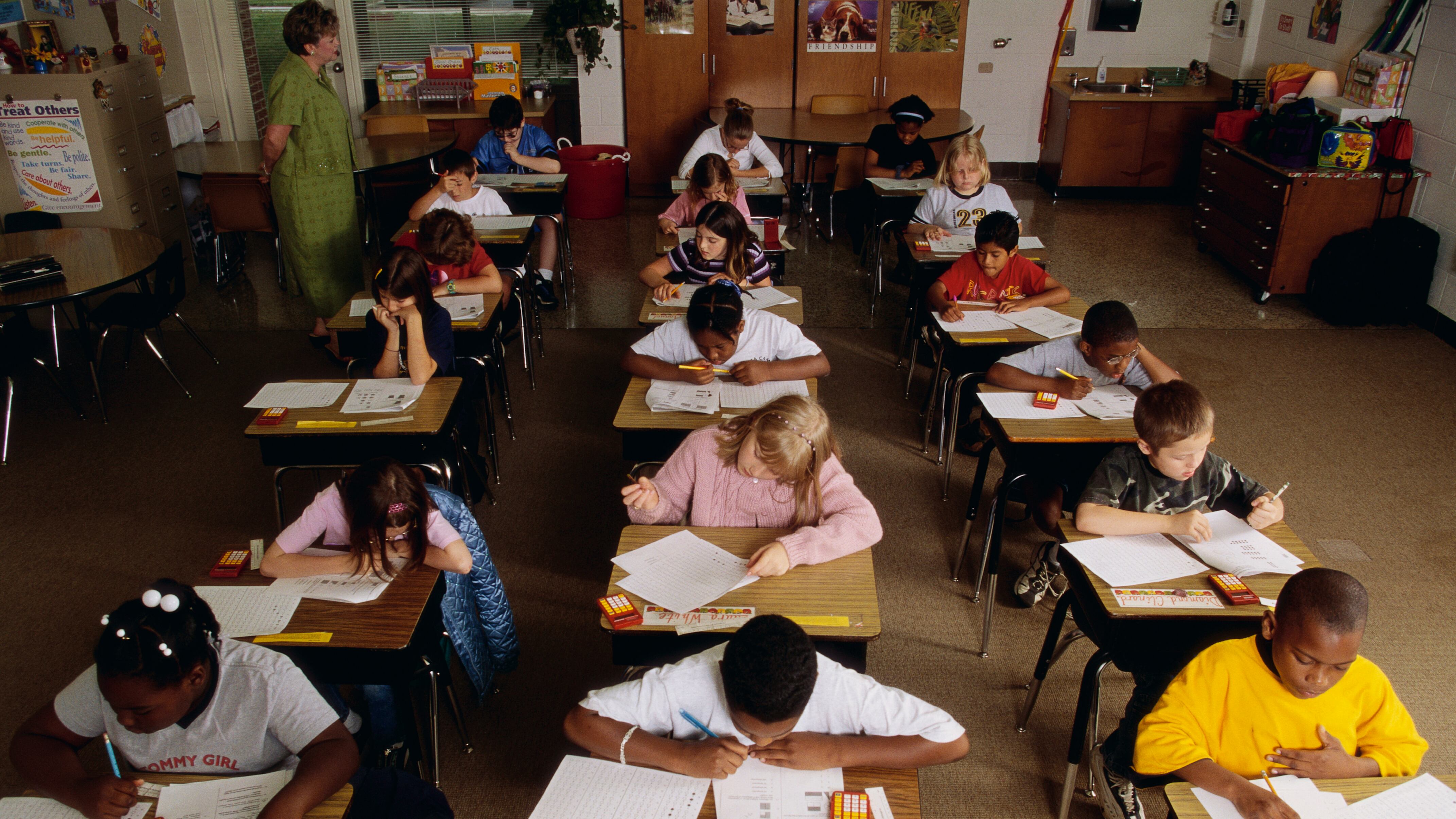 Eighteen elementary students take a standardized test at their desks while a teacher watches.