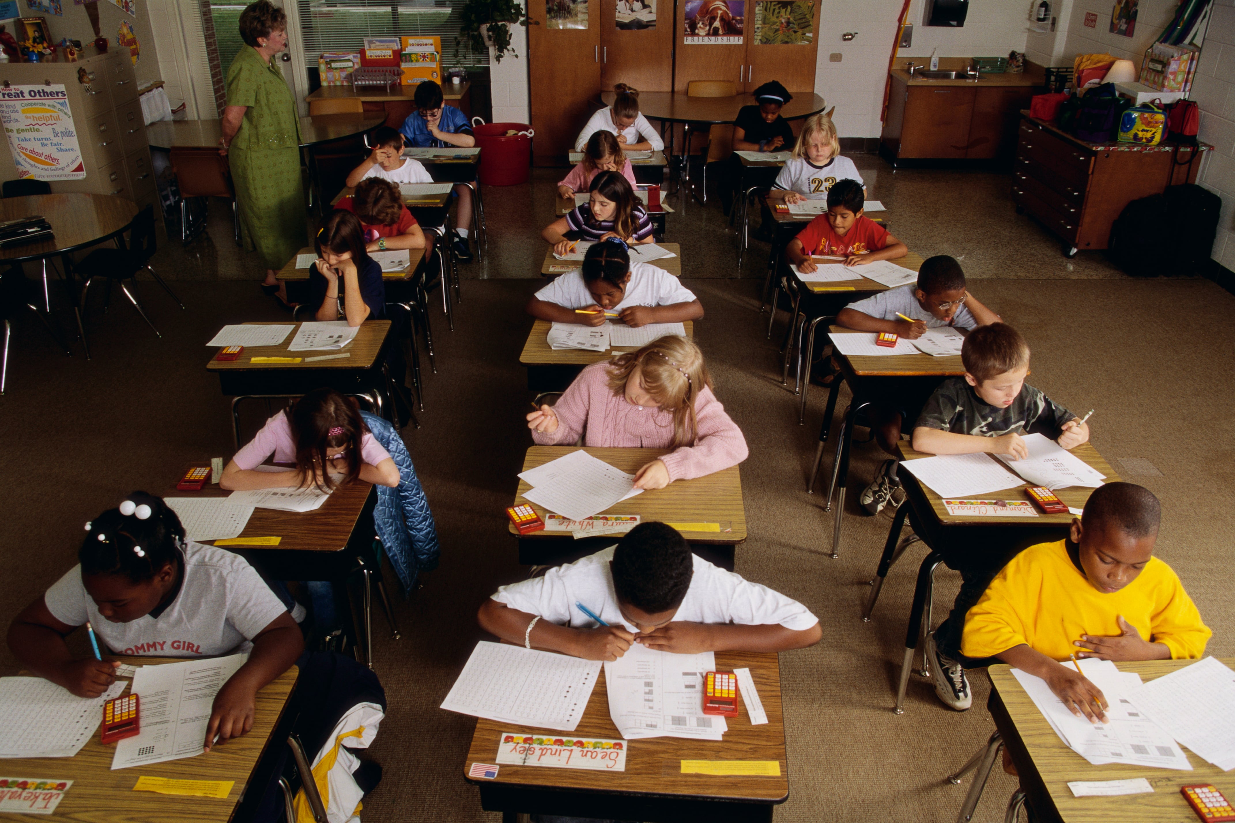 Eighteen elementary students take a standardized test at their desks while a teacher watches.