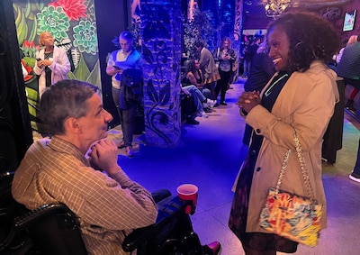A woman with a light jacket and dark dress talks to a man in a wheelchair with a checked shirt and jeans.
