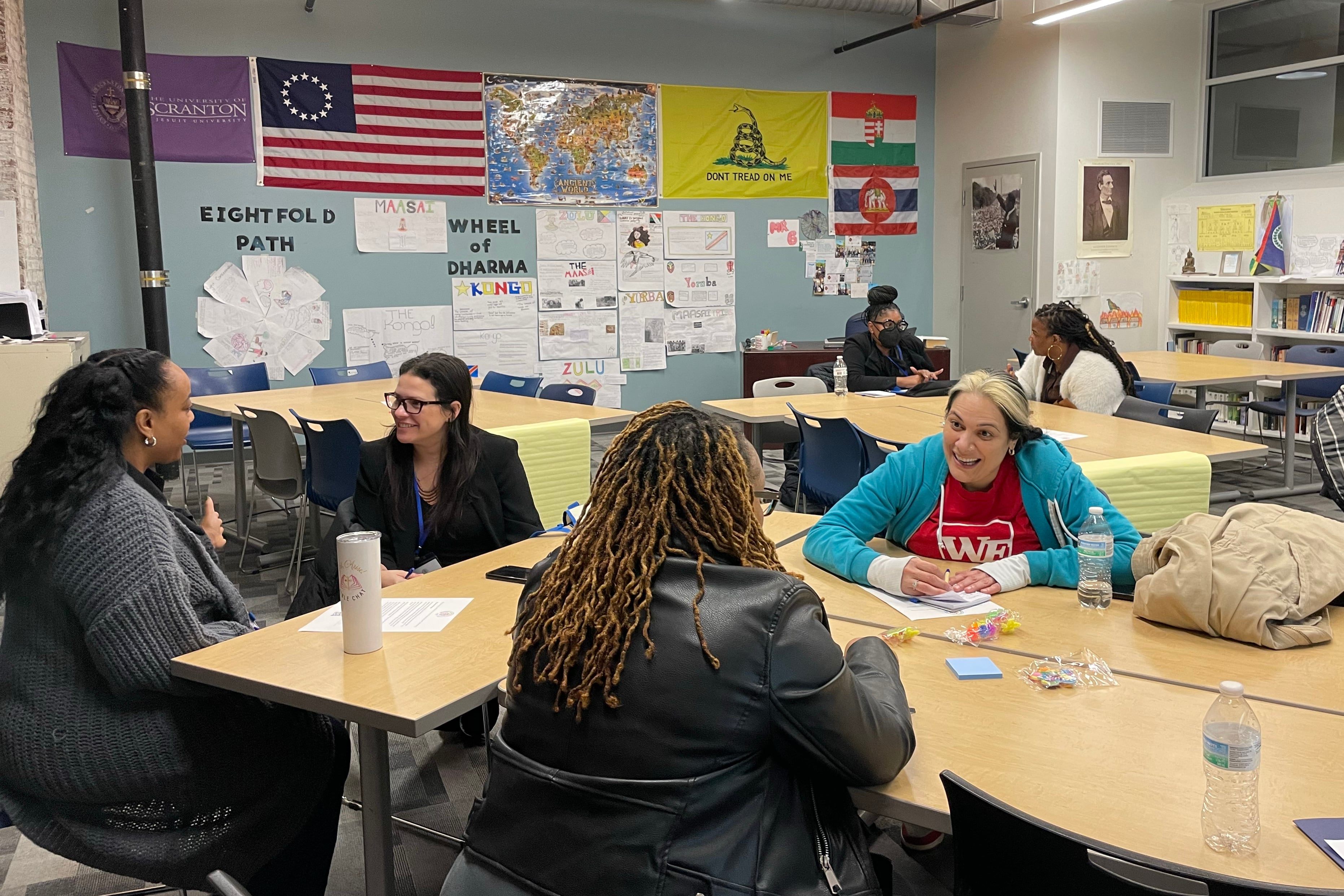 Four adults sit at a table talking in a classroom with a couple of people in the background.