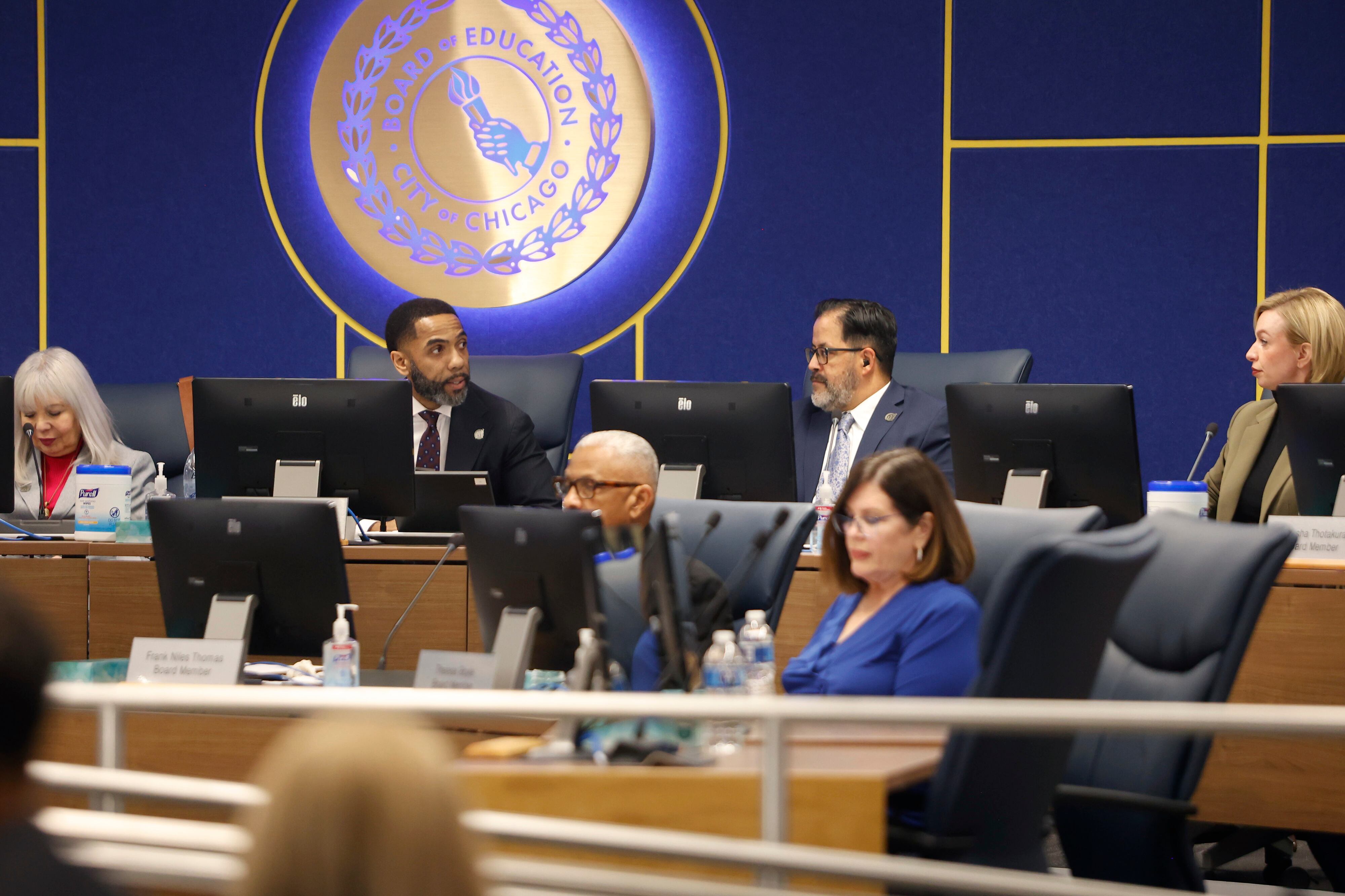 People in business clothes sit in a large conference room with a blue wall and an illuminated circular sign in the background.