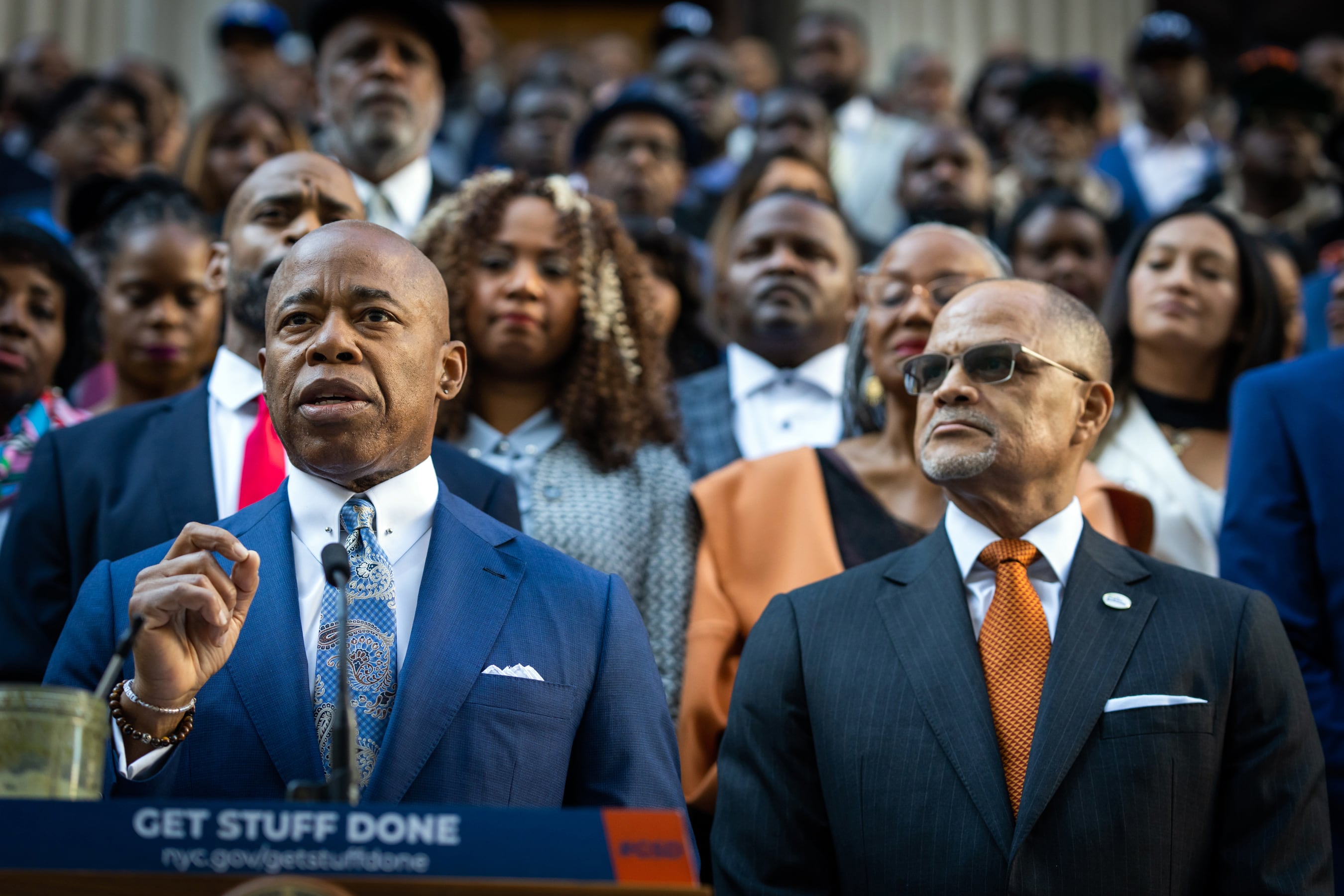 One man in a suit stands in front of a microphone with rows of people standing behind him.