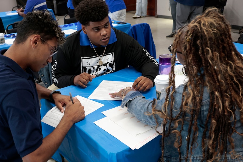 Three Black high school students sit at a table filling out voter registration paperwork in a room.