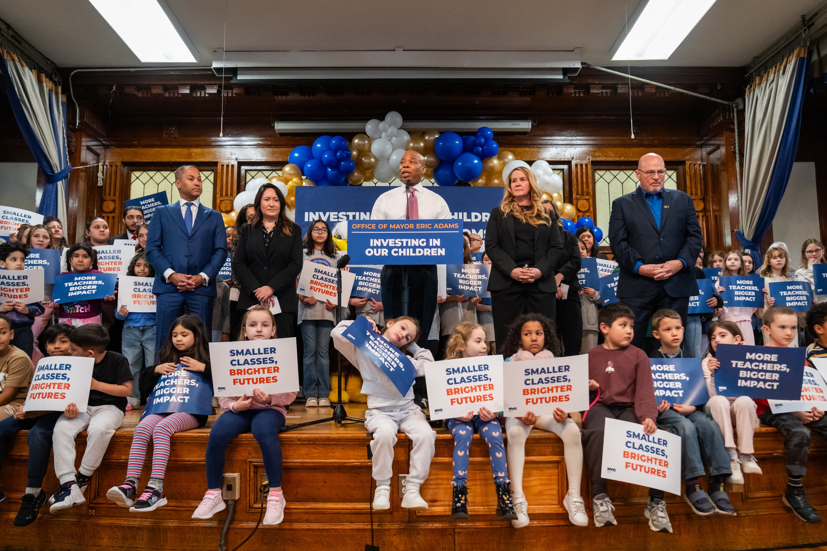 A group of officials in suits are joined on a stage with kids holding signs.