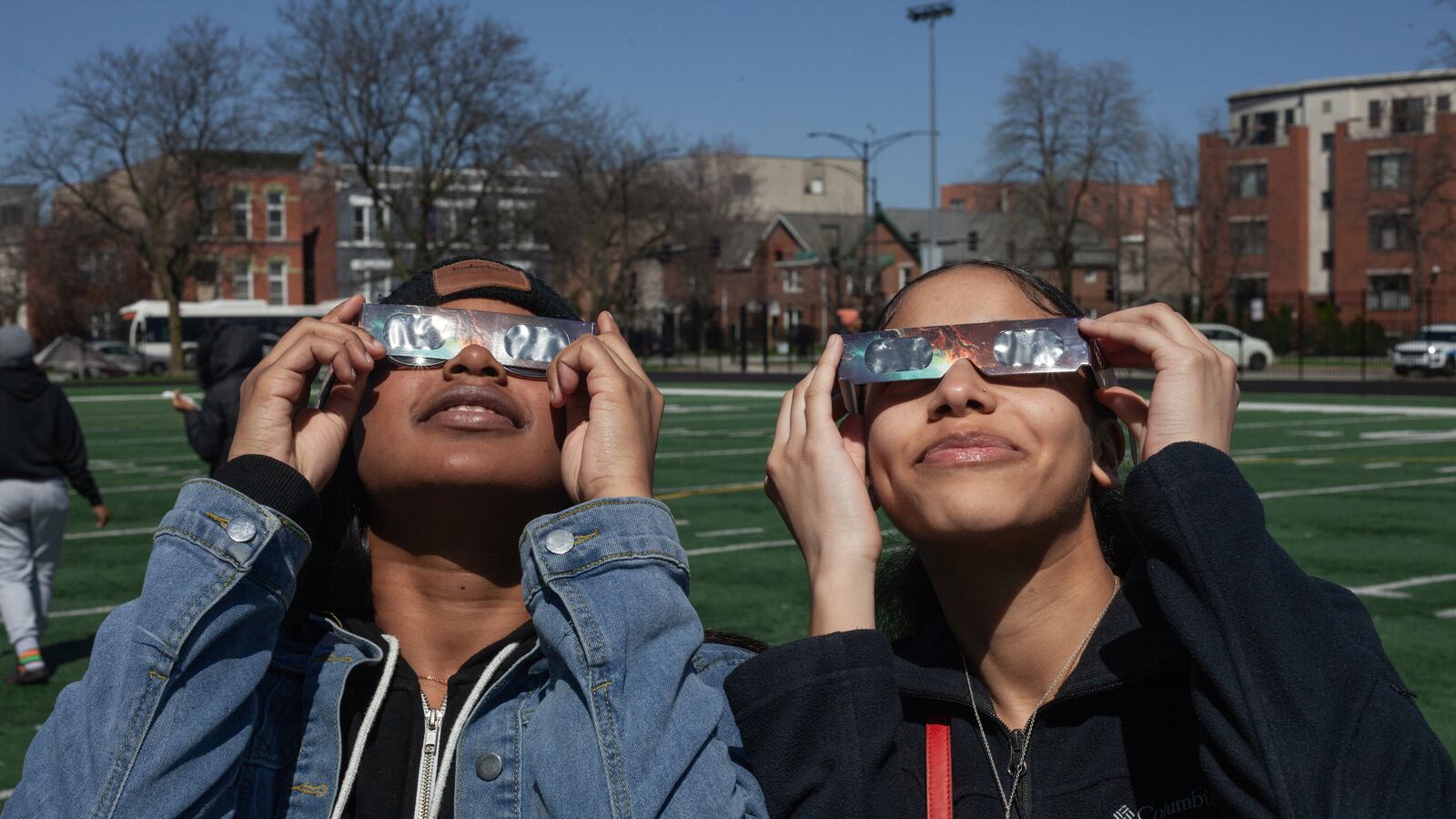 Two students holidng eclipse viewing glasses look up at the sky with a row of buidlings in the background.
