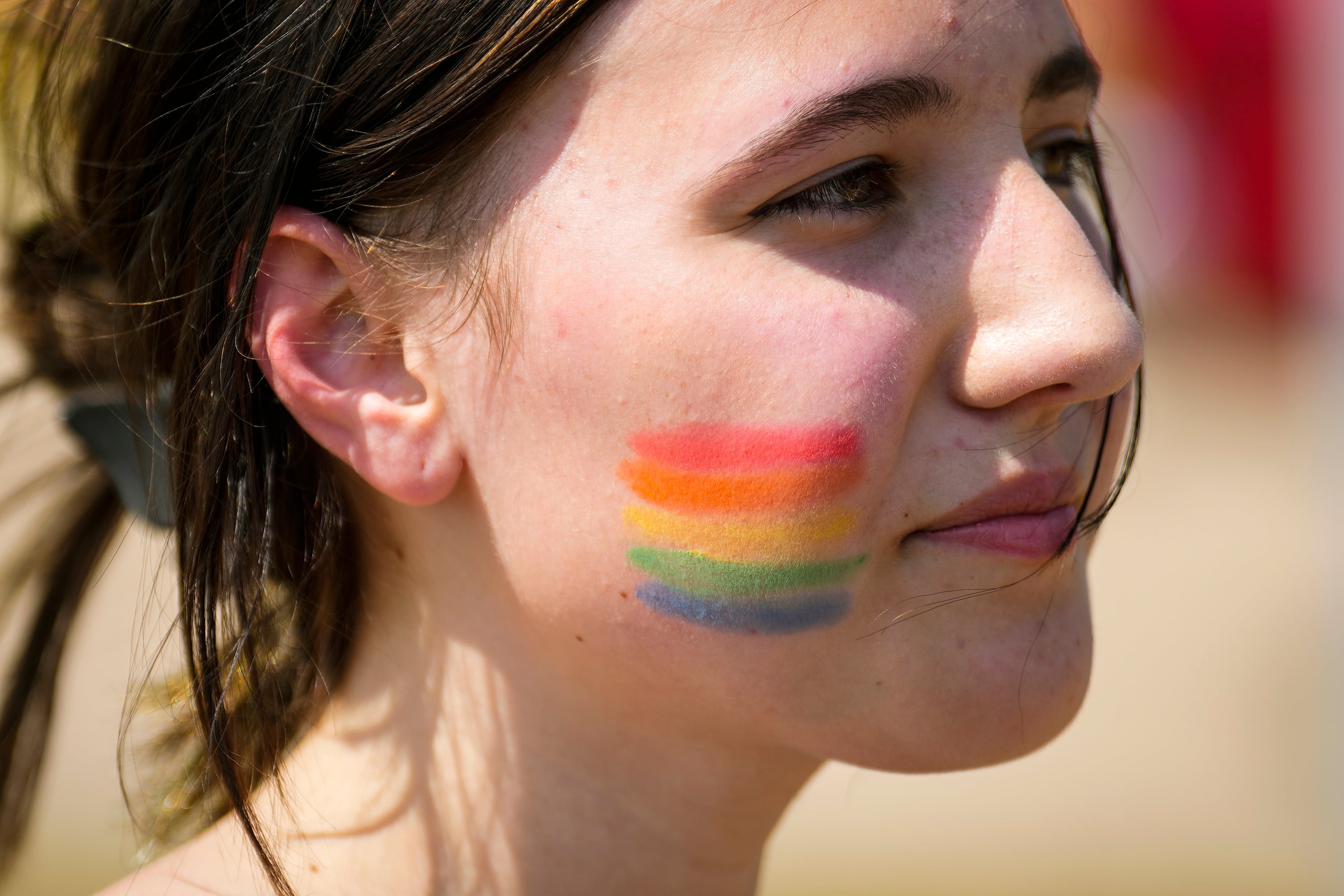 A close up of a young person's face with dark brown hair tied up and a rainbow of colors painted on their cheek.