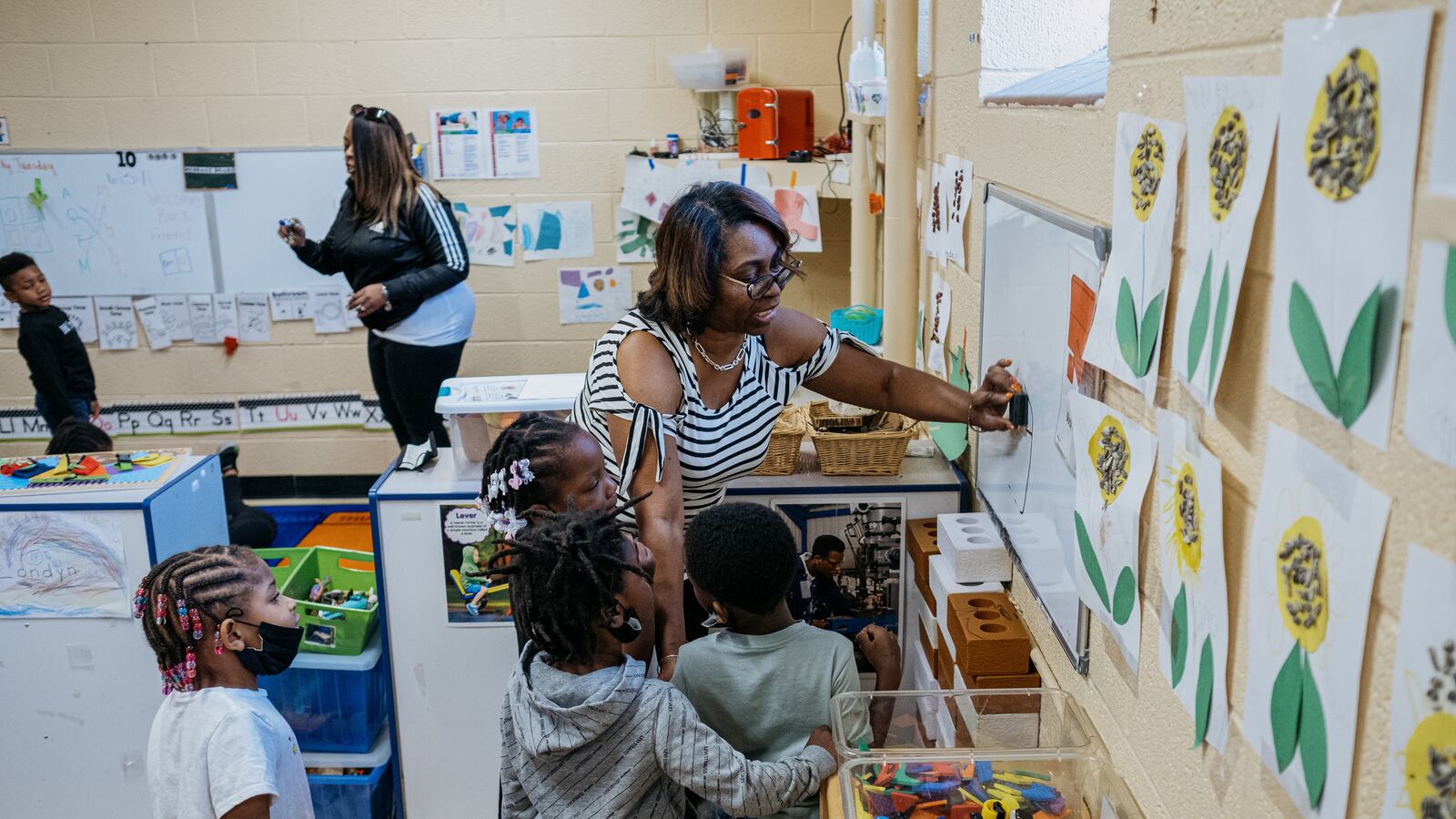 A teacher wearing glasses and a striped shirt speaks to a group of young students while they work at a whiteboard in a preschool classroom