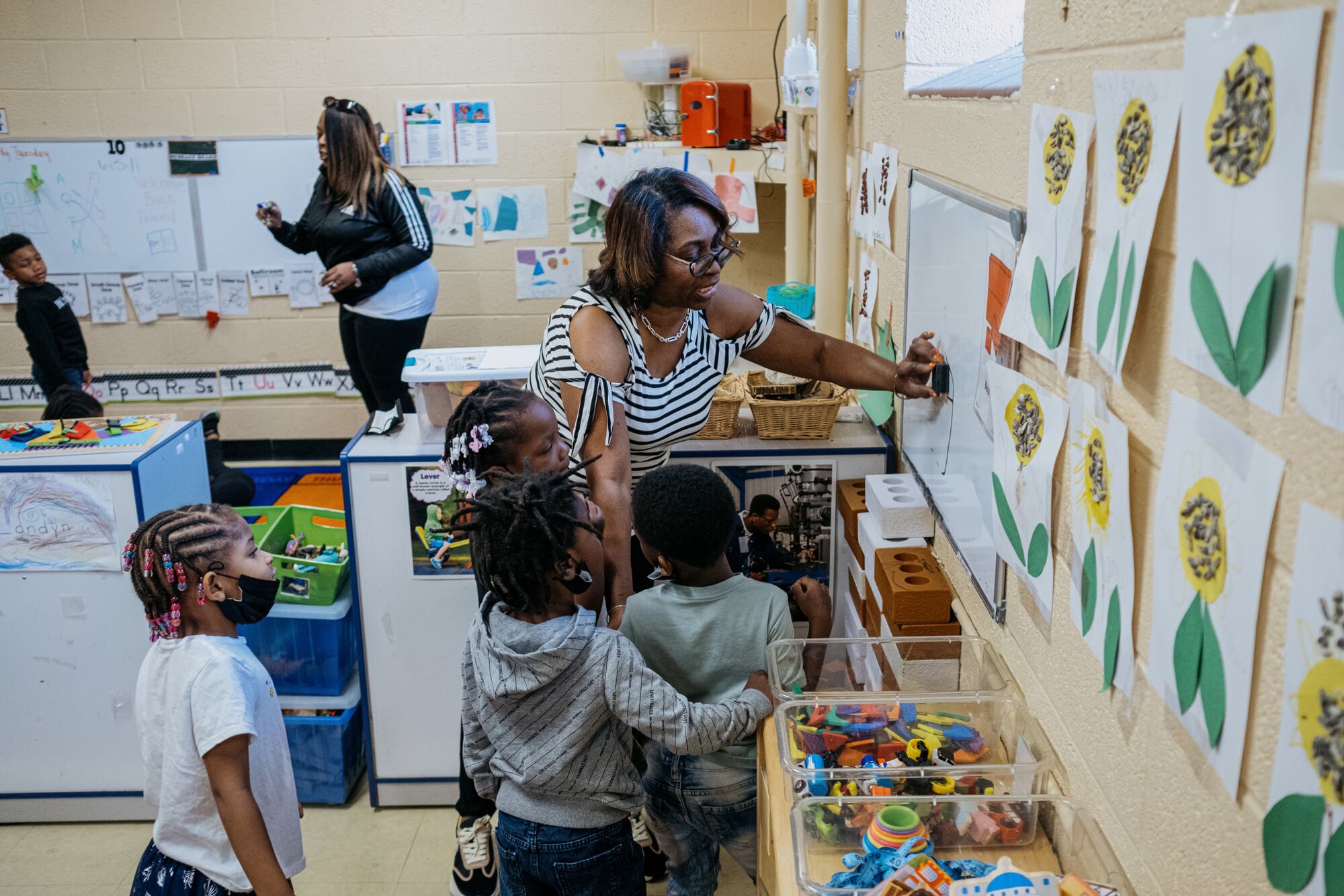A teacher wearing glasses and a striped shirt speaks to a group of young students while they work at a whiteboard in a preschool classroom