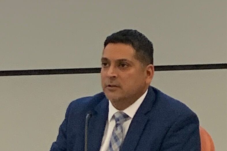Krish Mohip sits at a table and speaks into a microphone while wearing a blue suit and white shirt. A notepad is in front of him.