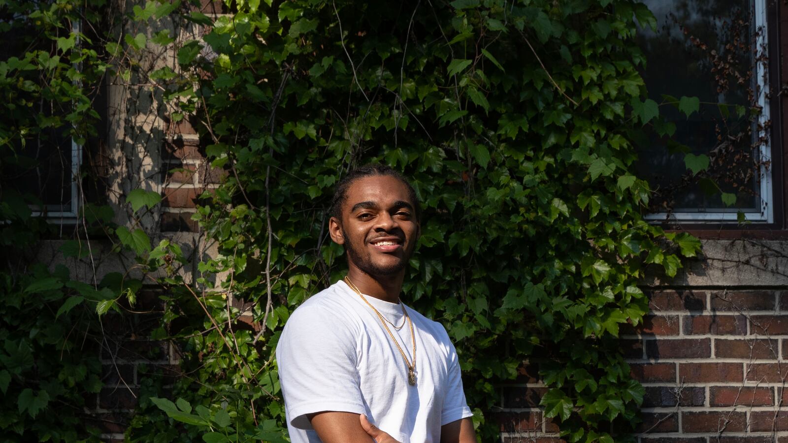 A high school senior with short dark hair and wearing a white t-shirt and a gold necklace with a diamond letter "K" crosses his arms and poses for a portrait while standing in front of a brick wall with greenery crawling up the wall.