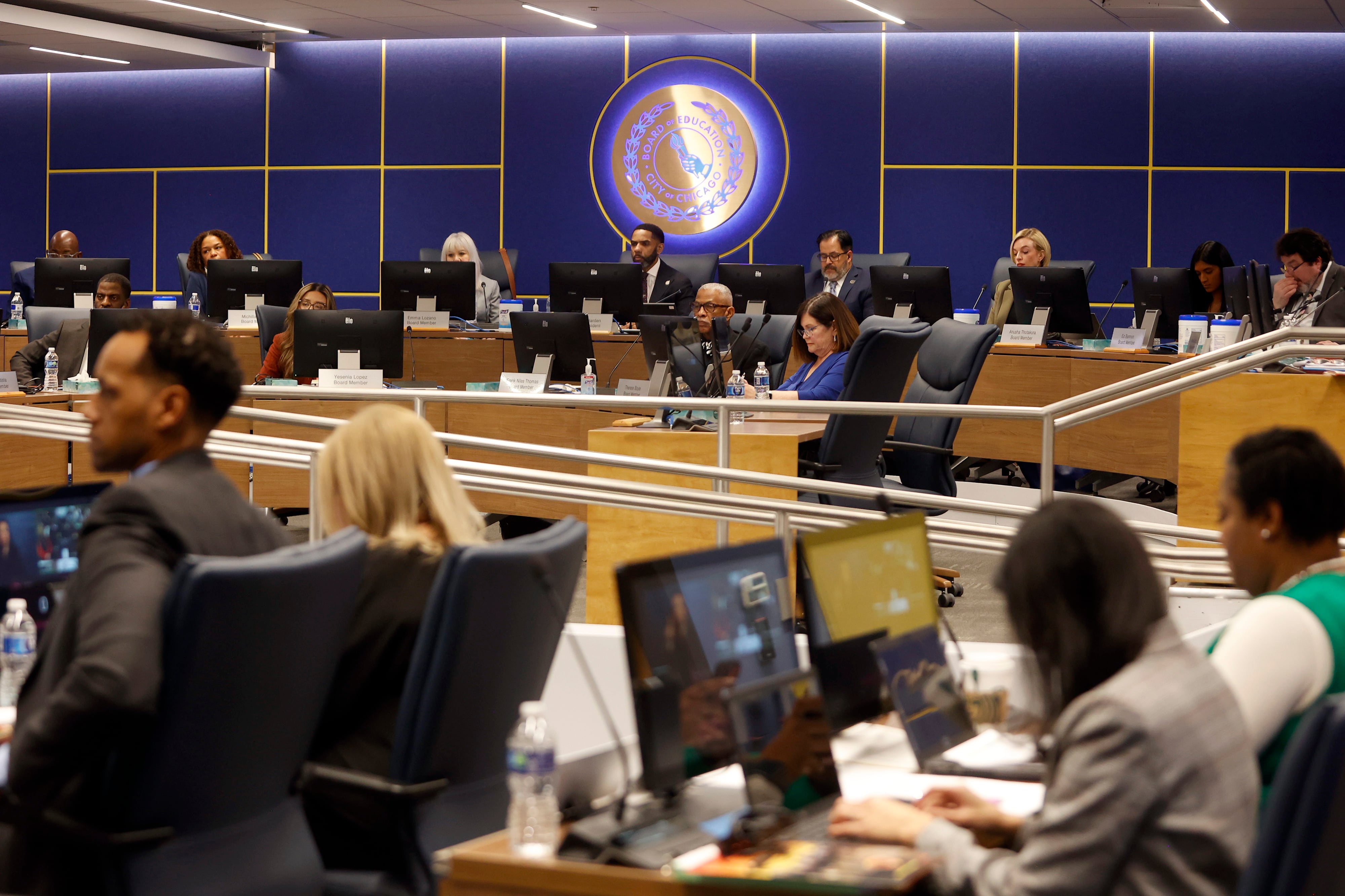People in business clothes sit in a large conference room with a blue wall and an illuminated circular sign in the background.
