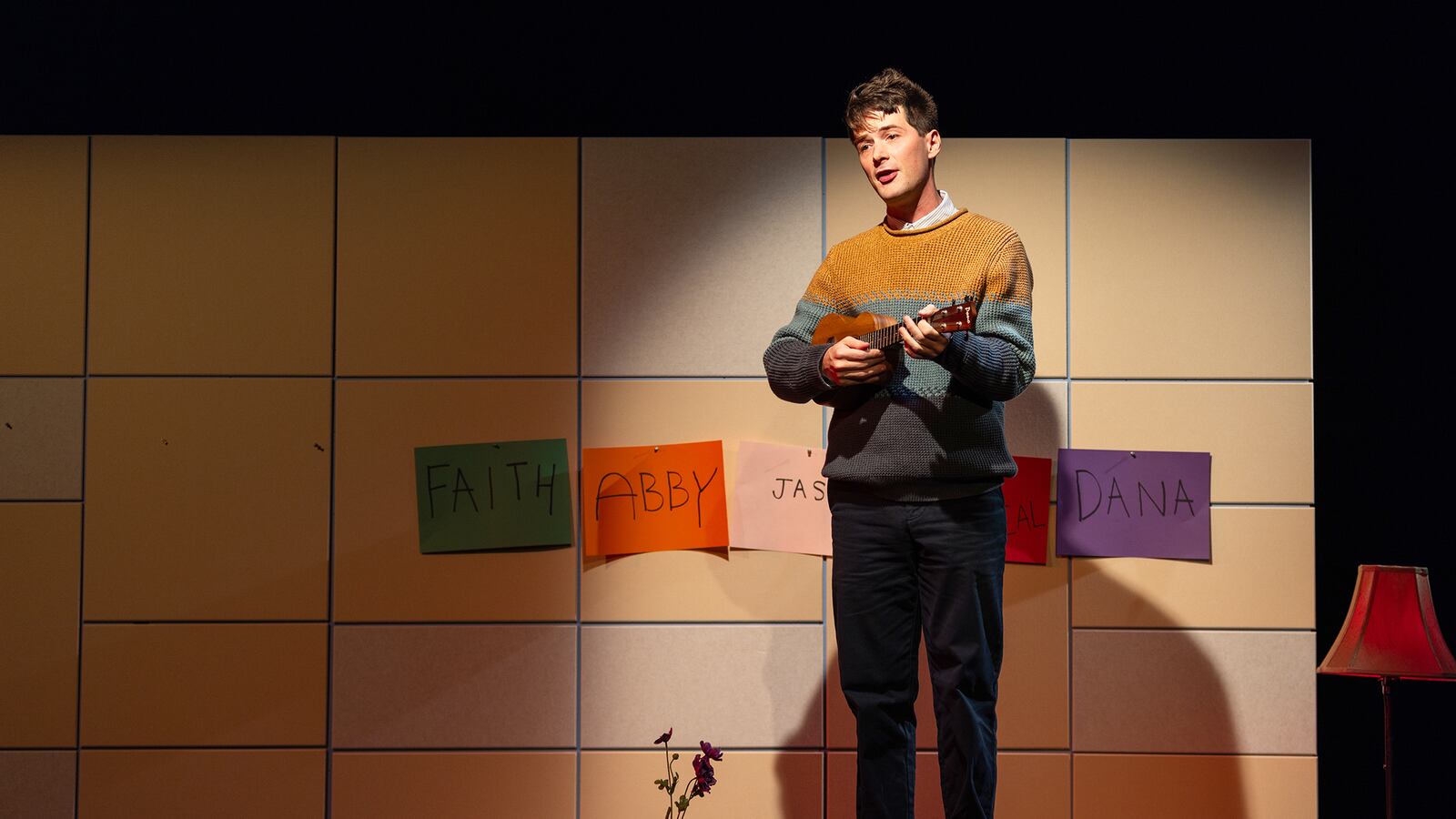 A person with short hair and wearing a multicolored sweater performs on a stage playing a ukulele.