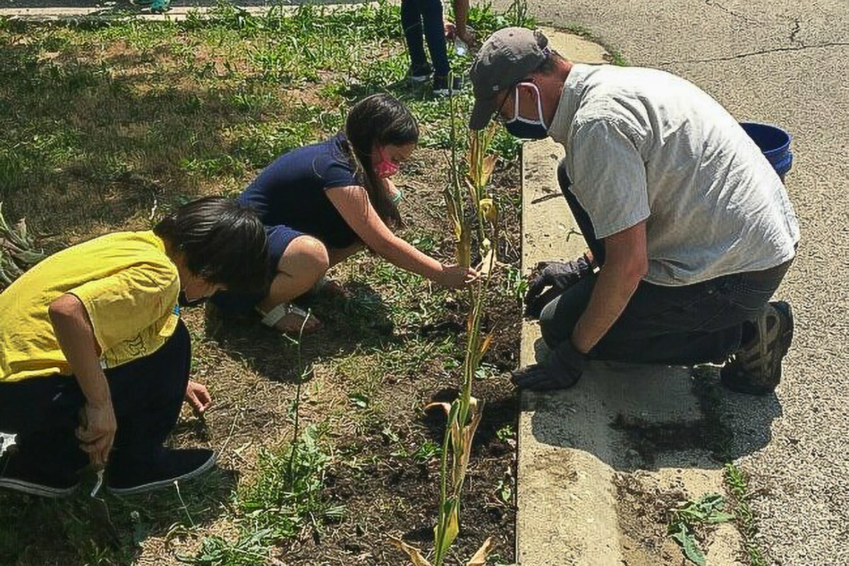 A male teacher wearing a grey hat plants on the side of a concrete curb with three students.