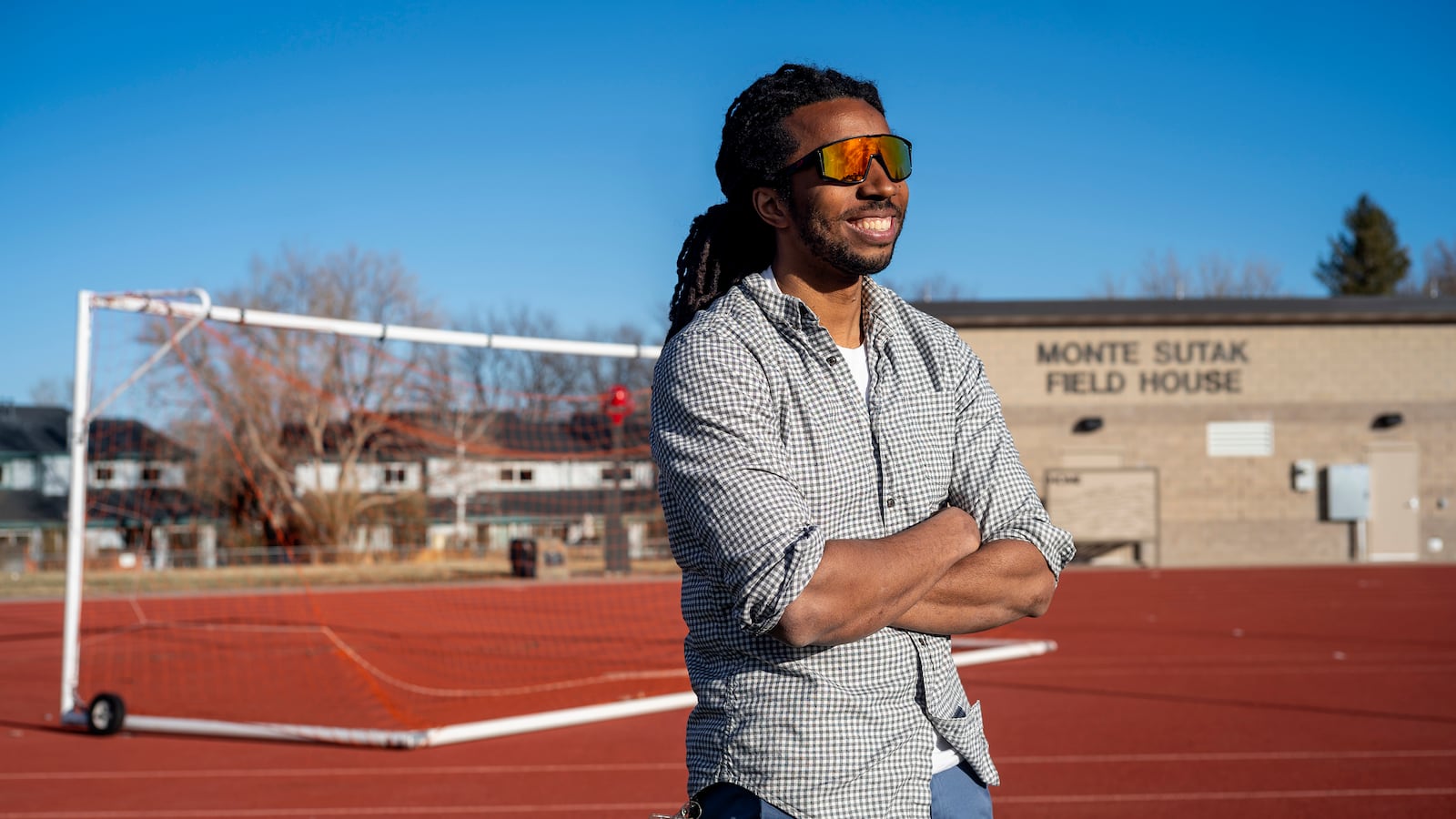 A young, Black educator and track coach, wearing a button-up shirt, and bright multi-colored sunglasses, smiles as he stands on the edge of the track.