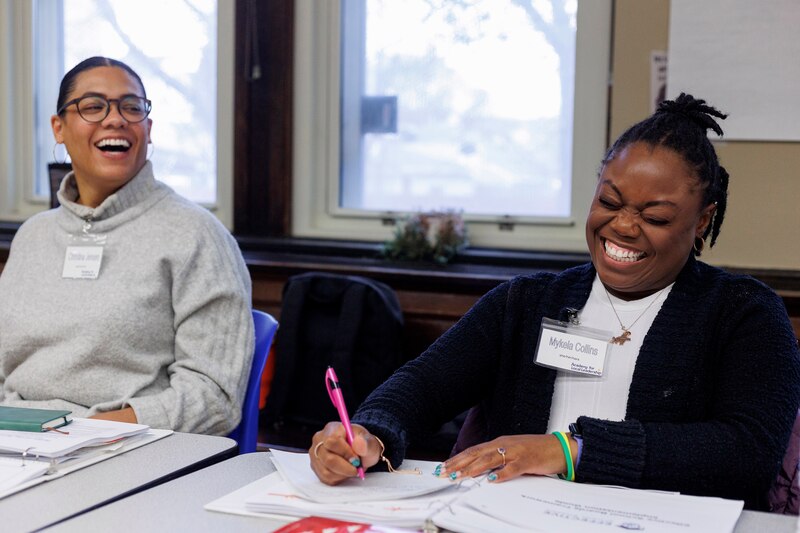 Two people laugh while sitting next to each other at a desk. The person on the right is writing on a piece of paper.