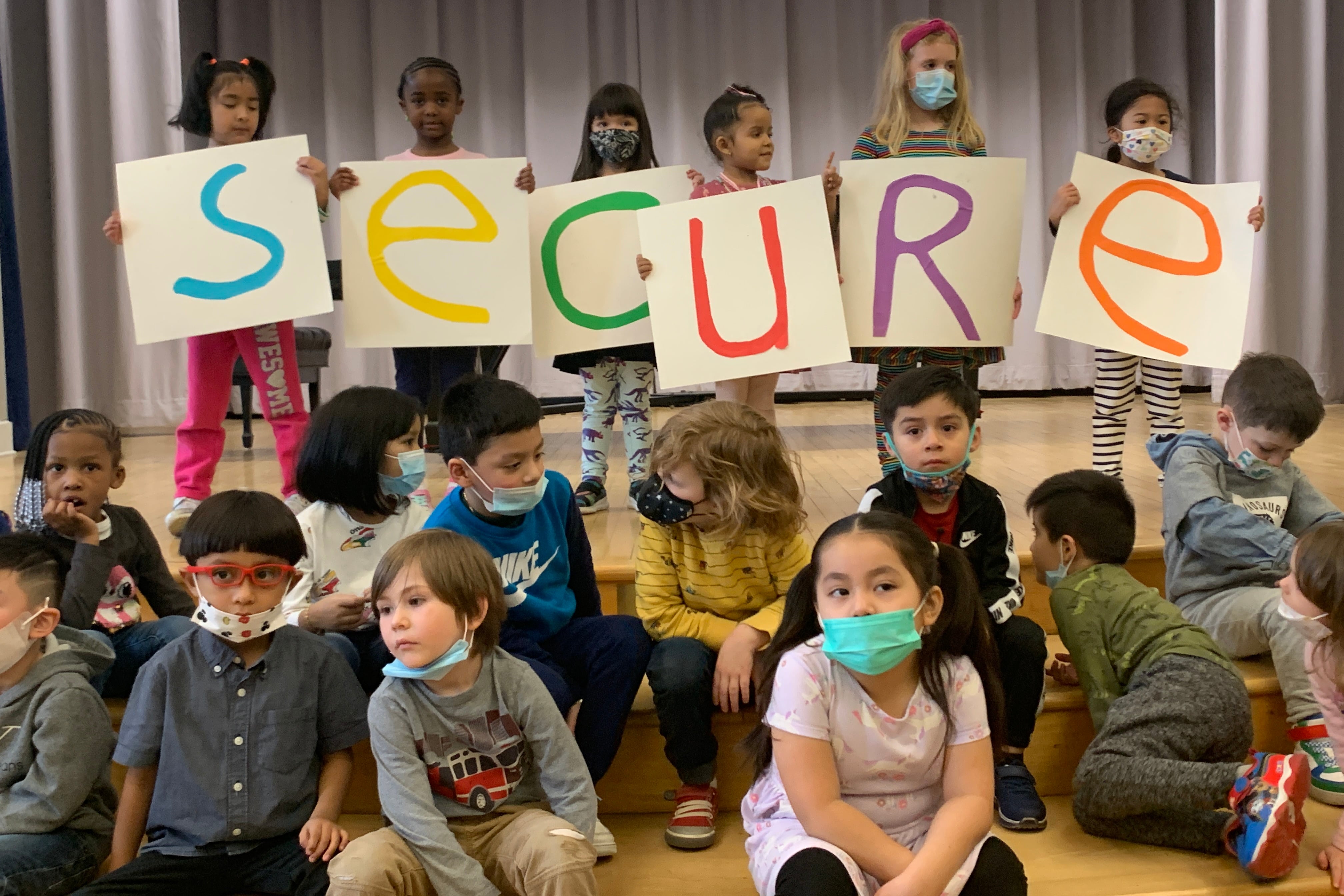 Several children hold up colored letters spelling out “Secure” while standing in front of a curtain as several other children sit in front of them. 