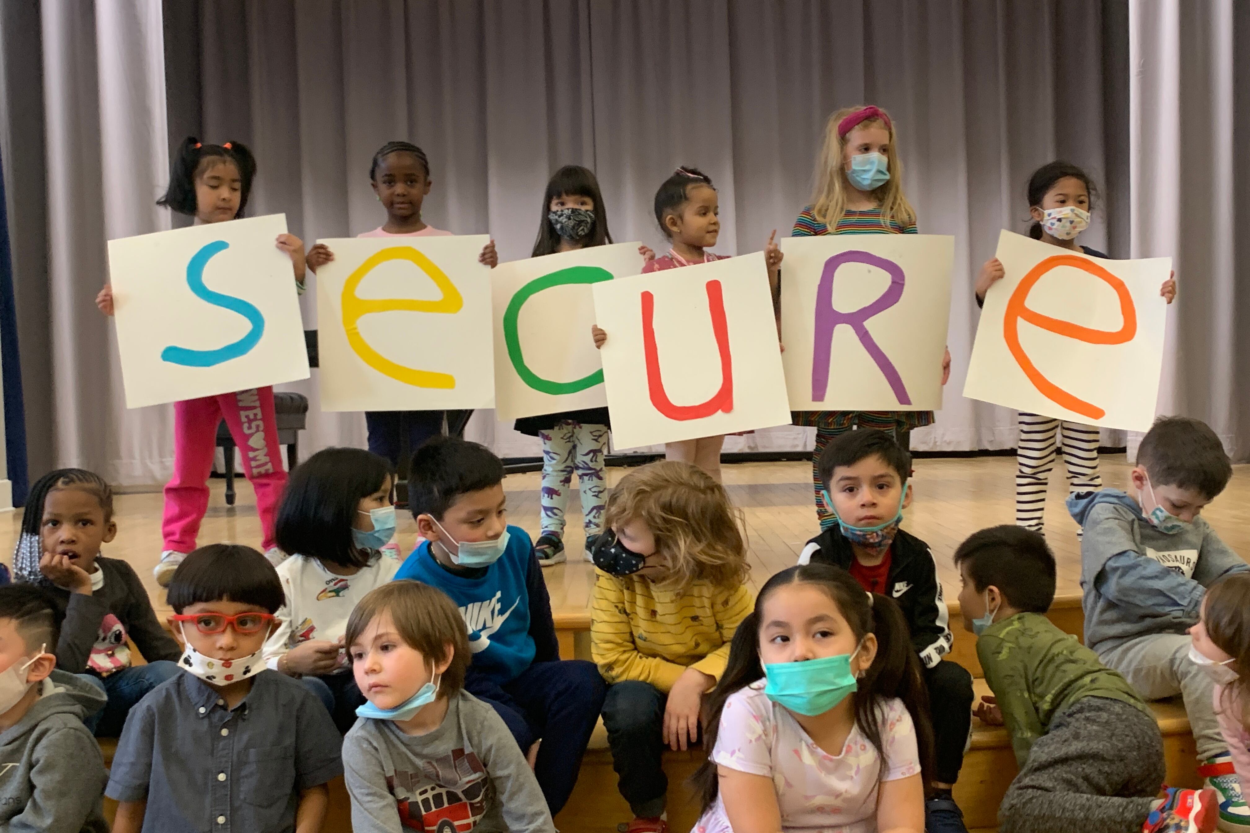 Several children hold up colored letters spelling out “Secure” while standing in front of a curtain as several other children sit in front of them.