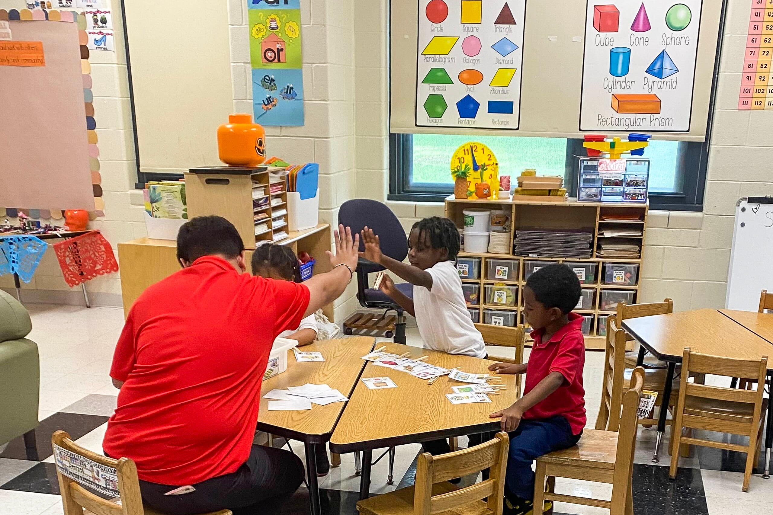 Three children sit around a table to work on an assignment in a colorful classroom. The educator, who is wearing a red shirt, gives a high five to a young student who is wearing a white collared shirt.