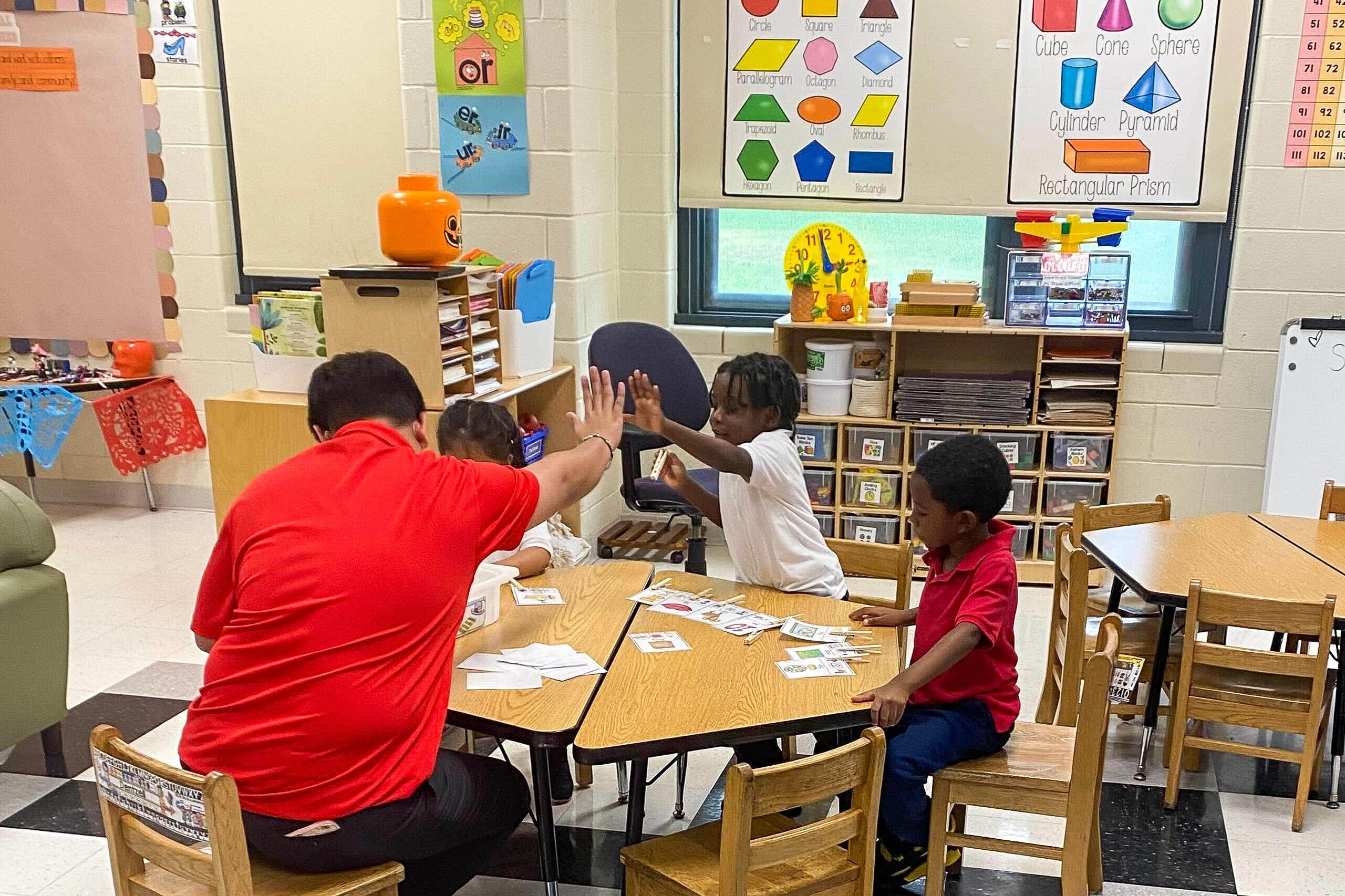 Three children sit around a table to work on an assignment in a colorful classroom. The educator, who is wearing a red shirt, gives a high five to a young student who is wearing a white collared shirt. 