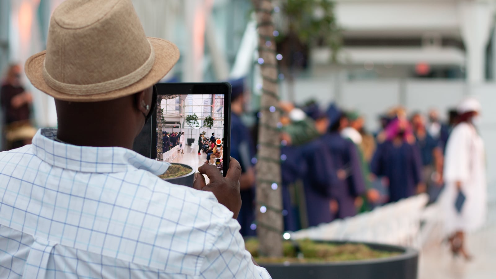 A parent wearing a straw hat and light checkered shirt uses an iPad to document graduation.