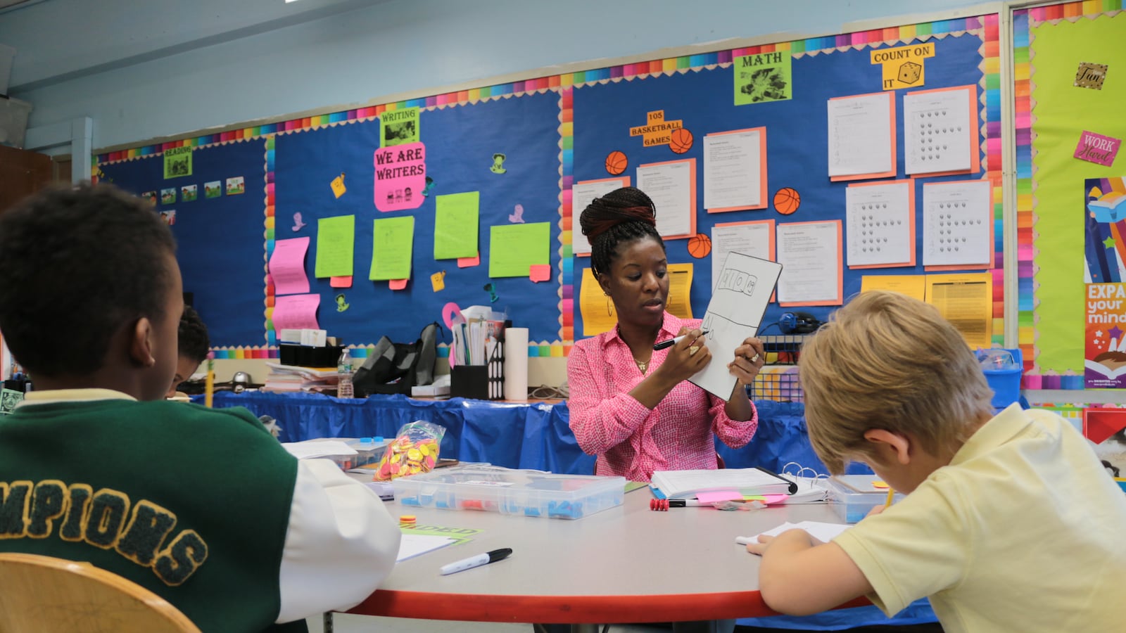 Two students work at a table, facing away from the camera, while a teacher in a pink shirt on the other side of the table writes on a small hand-held white board.