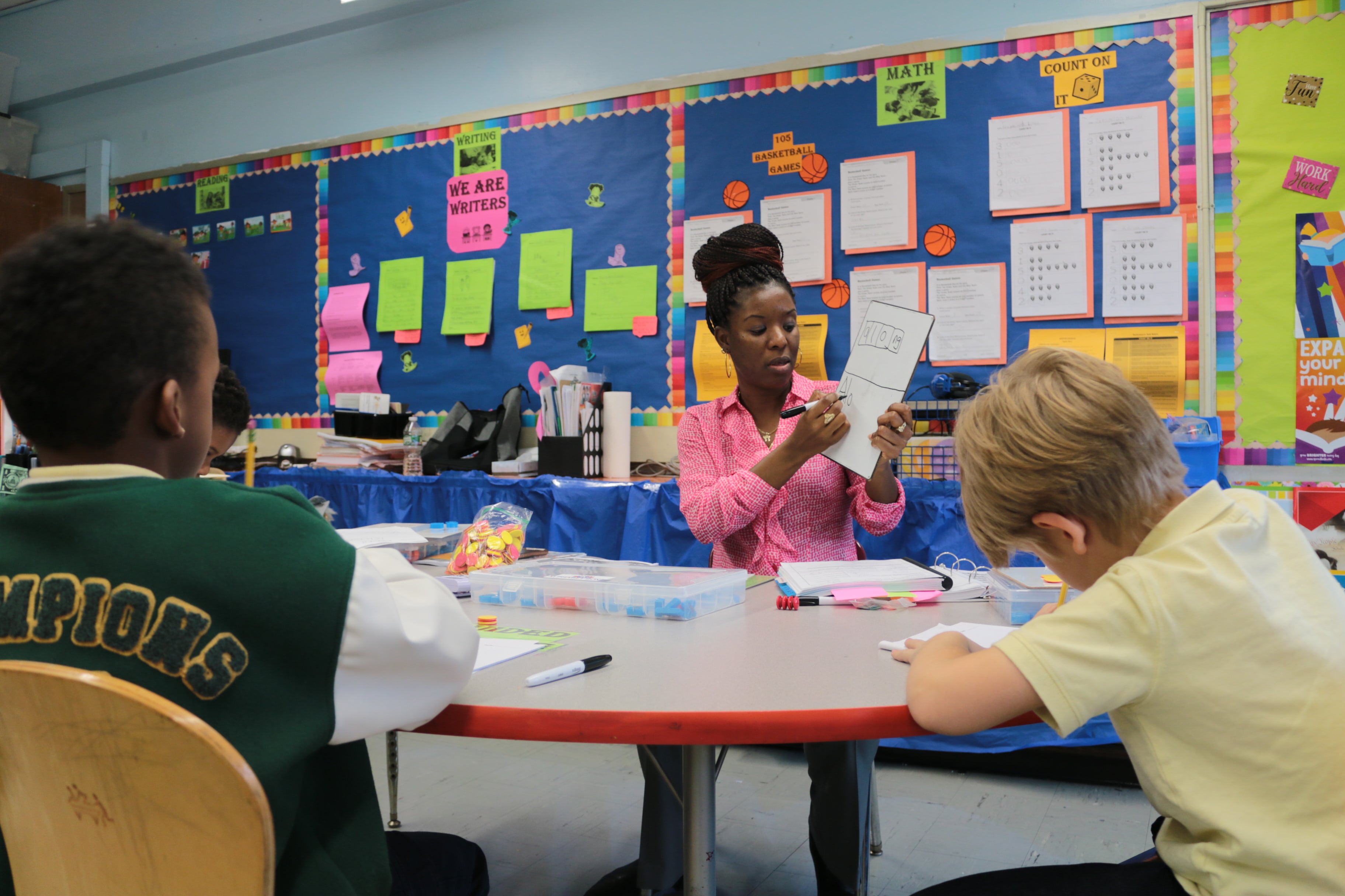 Two students work at a table, facing away from the camera, while a teacher in a pink shirt on the other side of the table writes on a small hand-held white board.