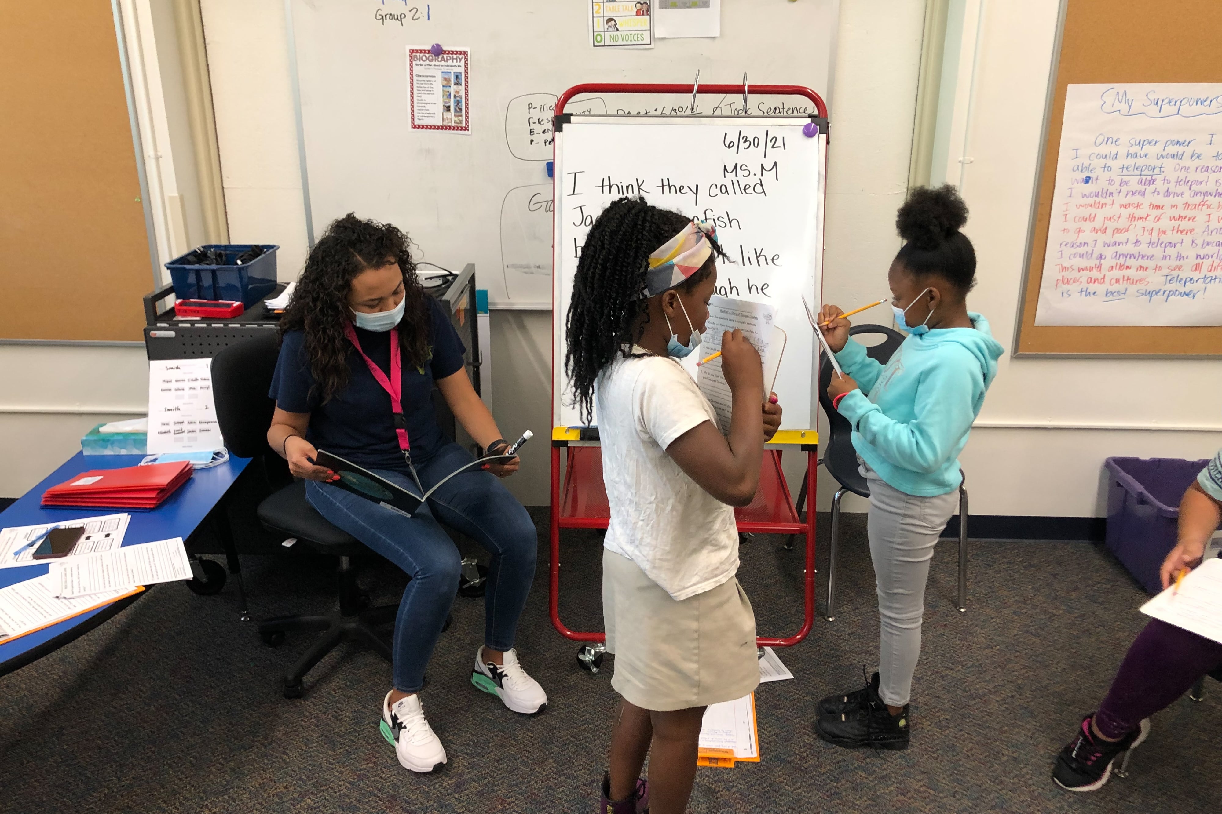 Two young girls work on reading comprehension in front of a small white board as their teacher sits next to them in the classroom. They are all wearing protective masks.
